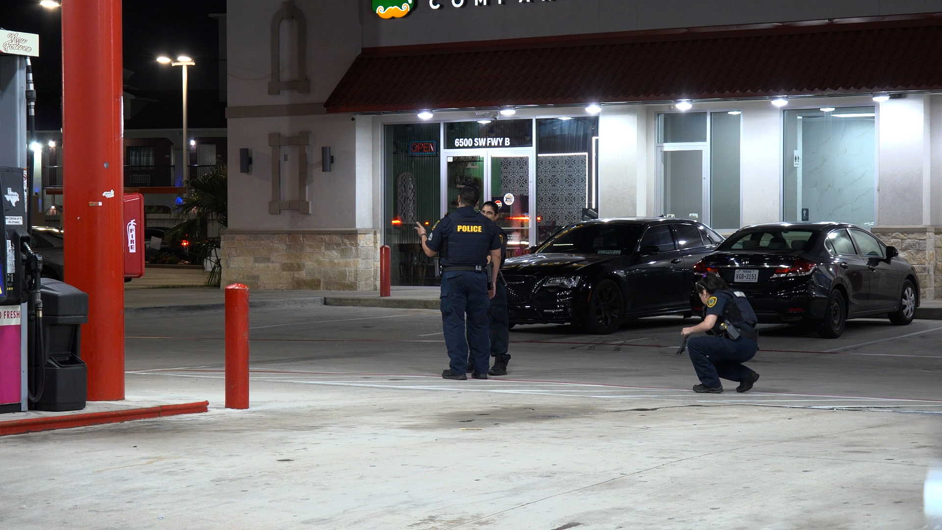 Nighttime scene at a gas station plaza with police officers in dark uniforms near a storefront and two black cars parked in front of the building.