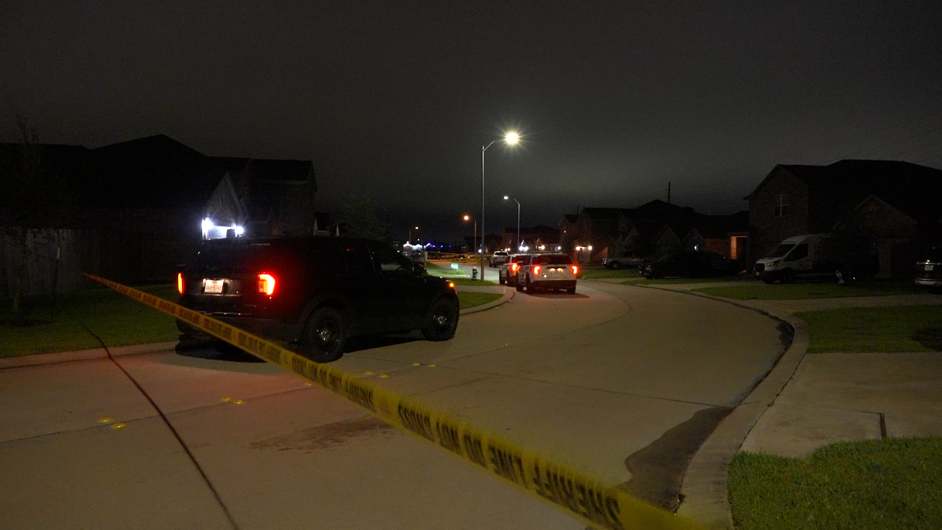Nighttime suburban street scene with yellow caution tape across the foreground and parked vehicles, including a black SUV, under streetlights and houses in the background.