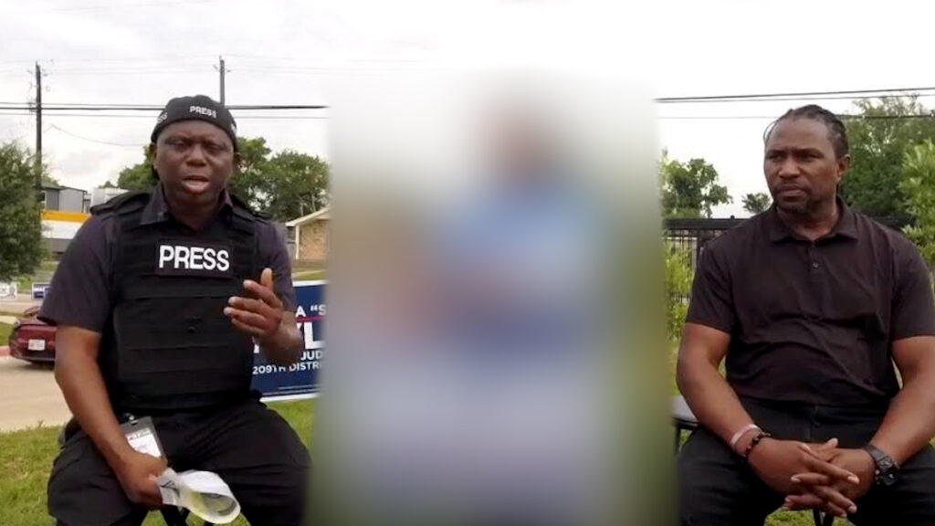Two men sit on a bench outdoors, left is a man in a PRESS vest speaking, right a man in a black polo listening, with a blurred figure in the center.