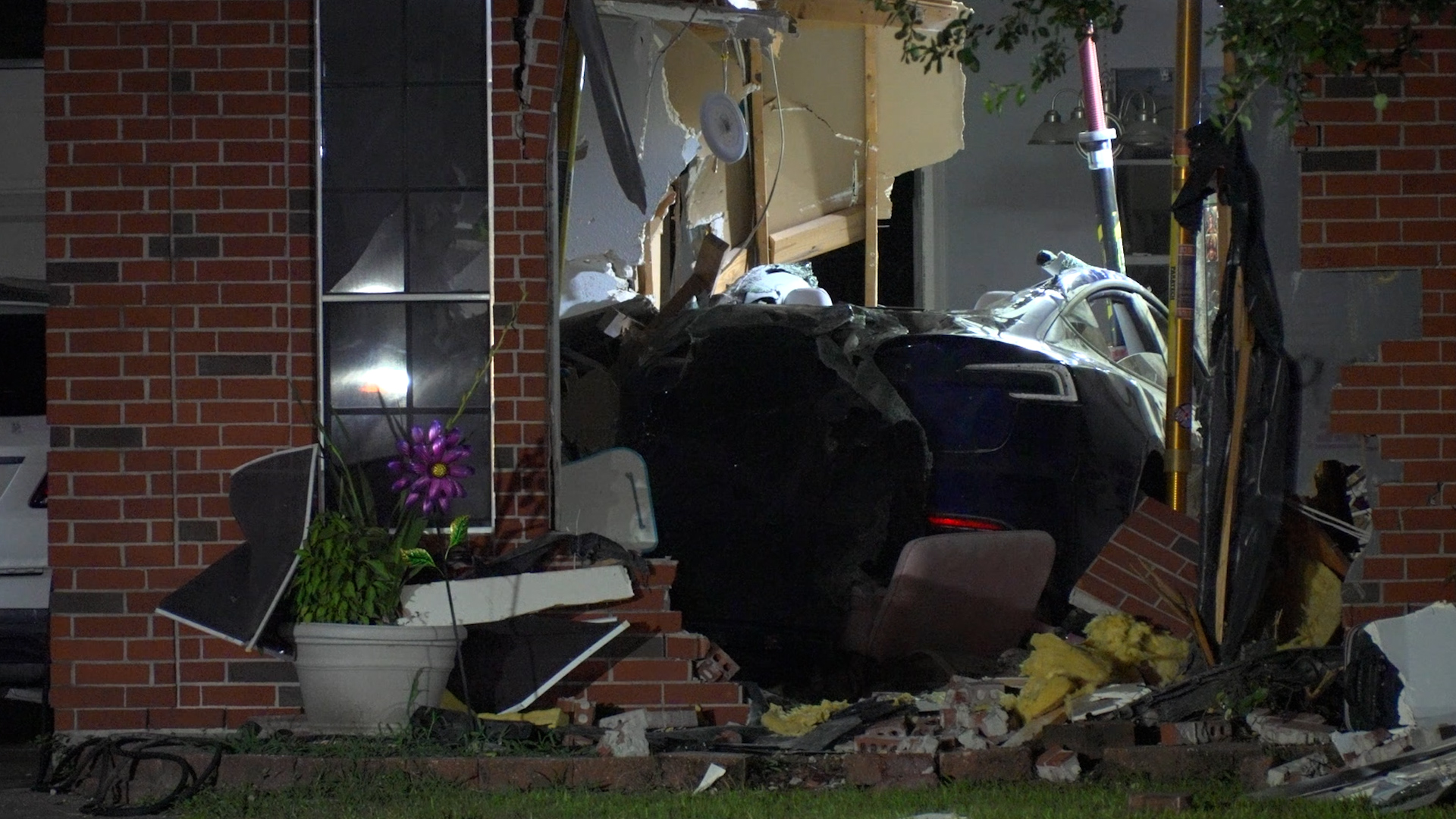 Car crashed into a brick house, with exposed interior and debris scattered outside the front.