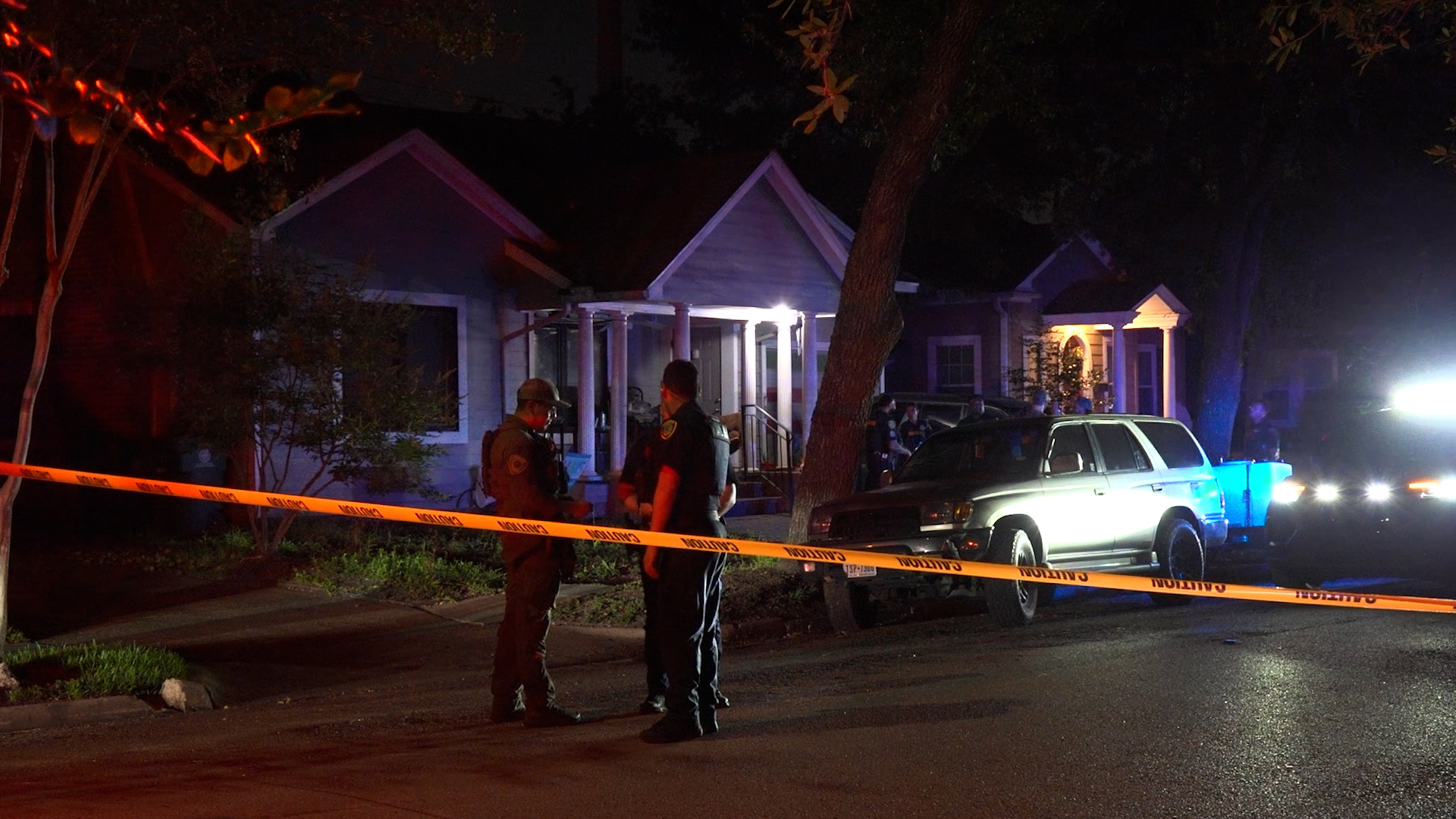 Nighttime crime scene on a residential street with police tape and officers standing near a gray SUV and a house with porch lights.