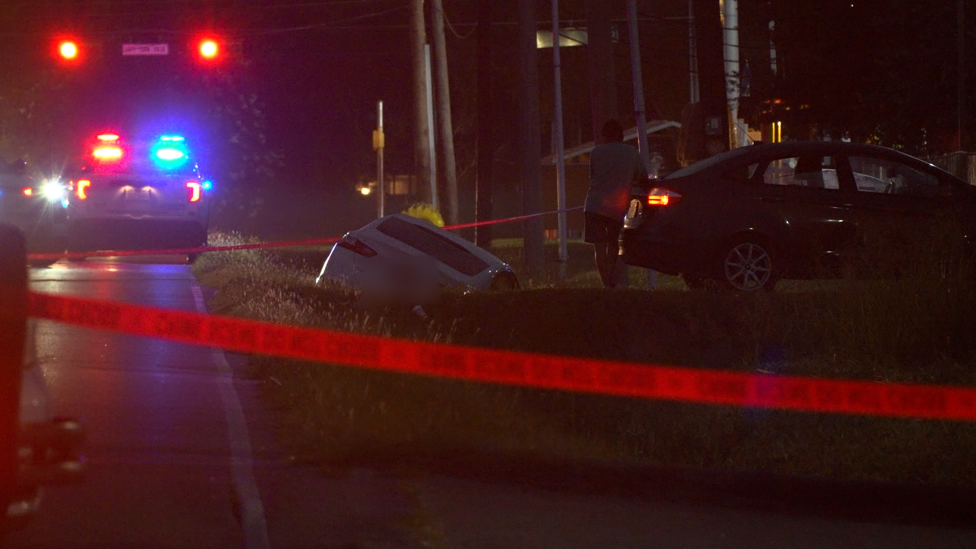 Nighttime car crash scene with a white car tipped on its side, a nearby black car with an open door, flashing police lights, and red caution tape nearby.