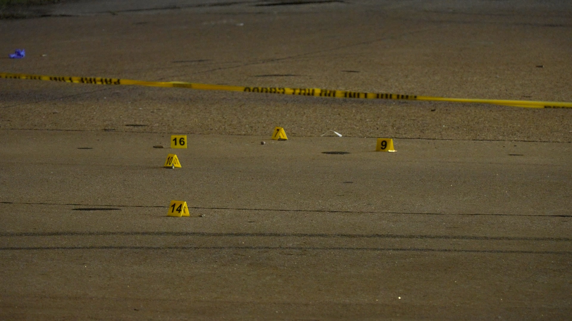 Yellow evidence markers with numbers scattered on a pavement, crime scene tape in the background.