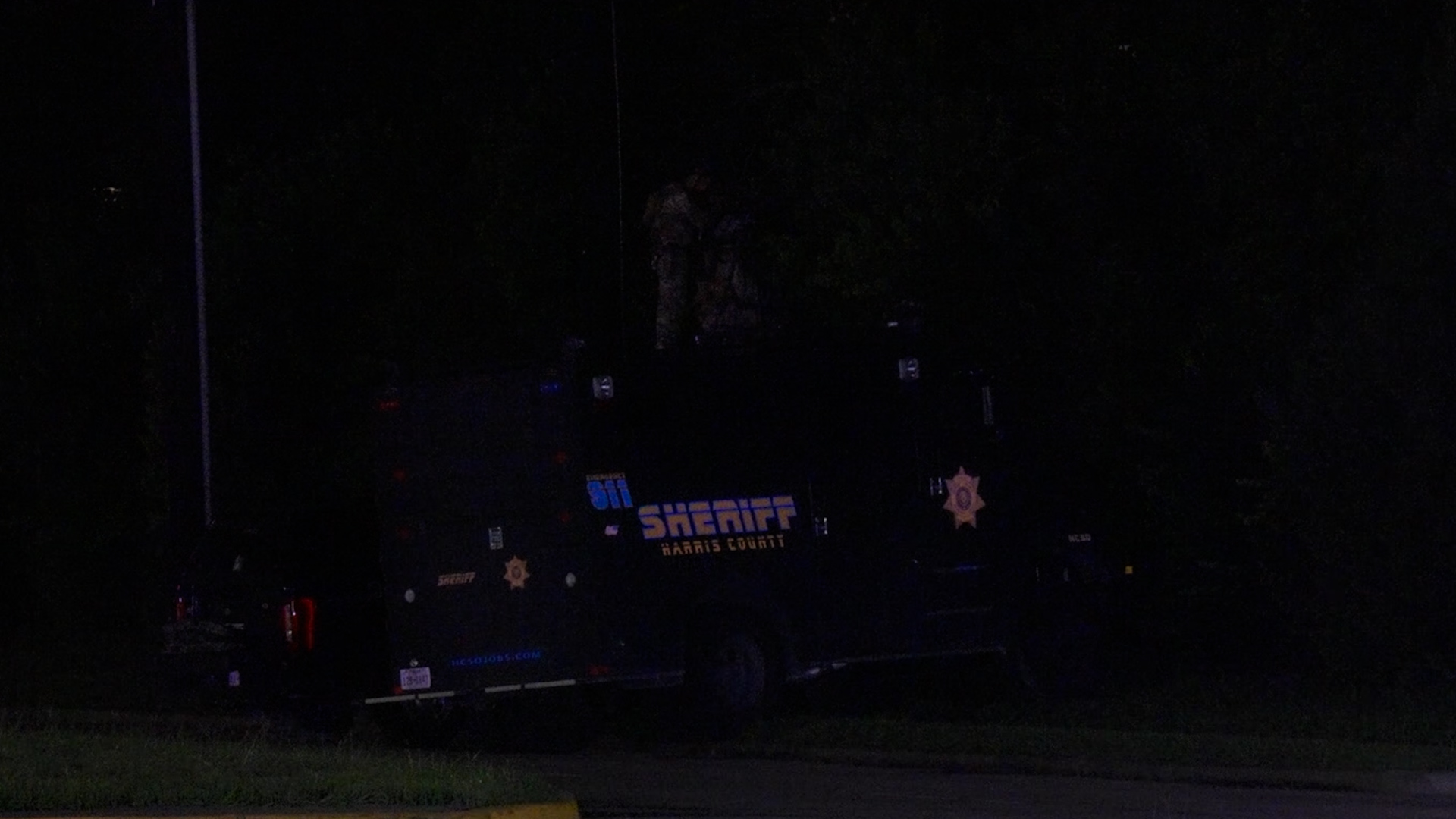 Dark nighttime scene showing a sheriff patrol vehicle with blue lights and a sheriff badge emblem on the door parked by the curb.