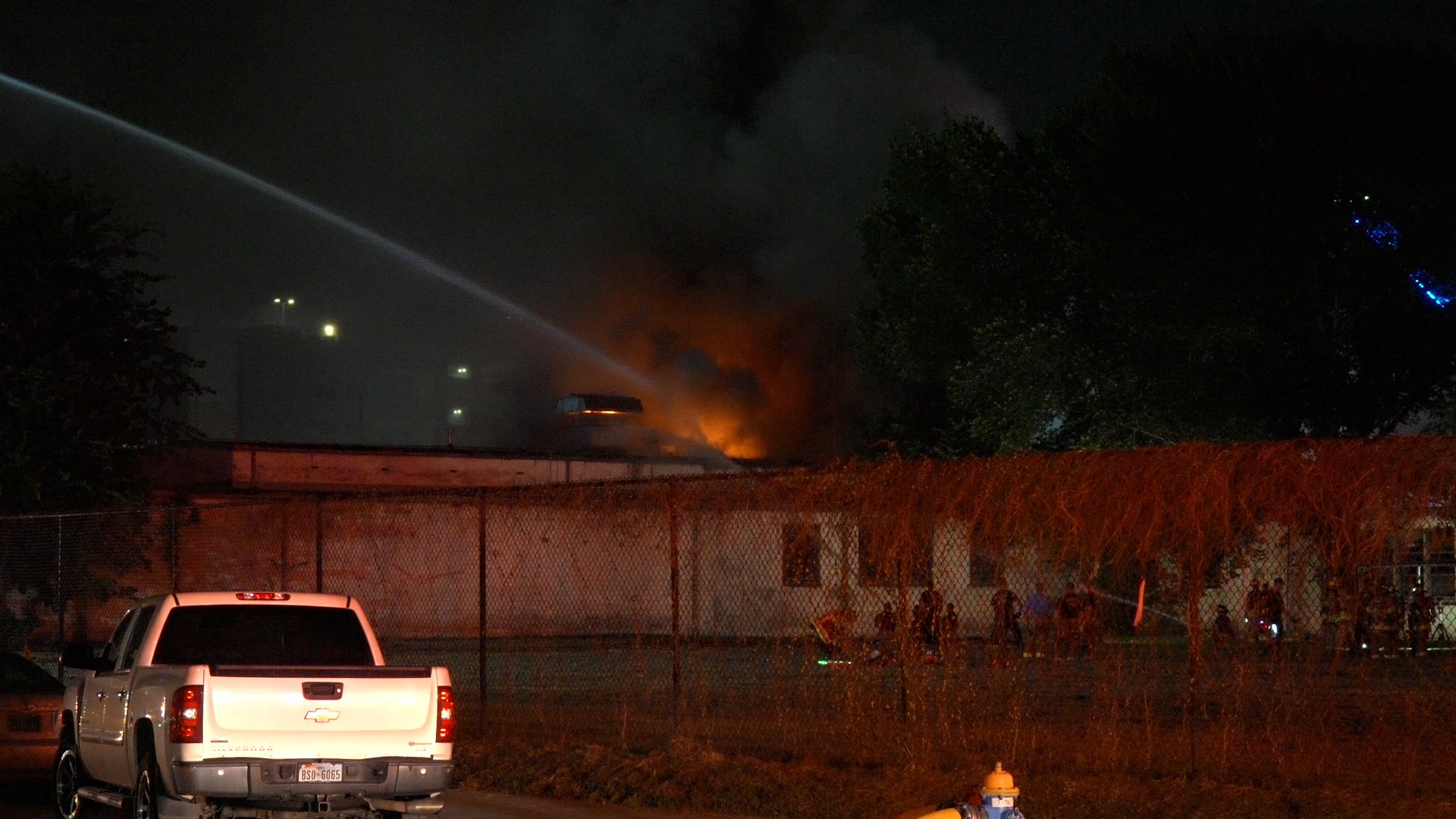 Nighttime exterior view of a vehicle on fire behind a chain-link fence, flames and dark smoke rising, with bystanders nearby.