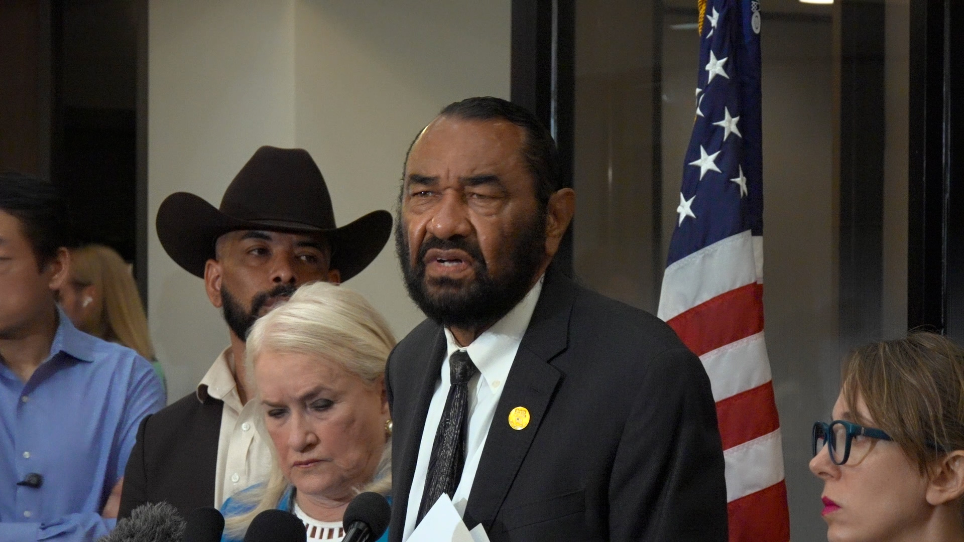 Man in a suit speaks at a press briefing with microphones in front, standing beside the U.S. flag as others listen in the background.
