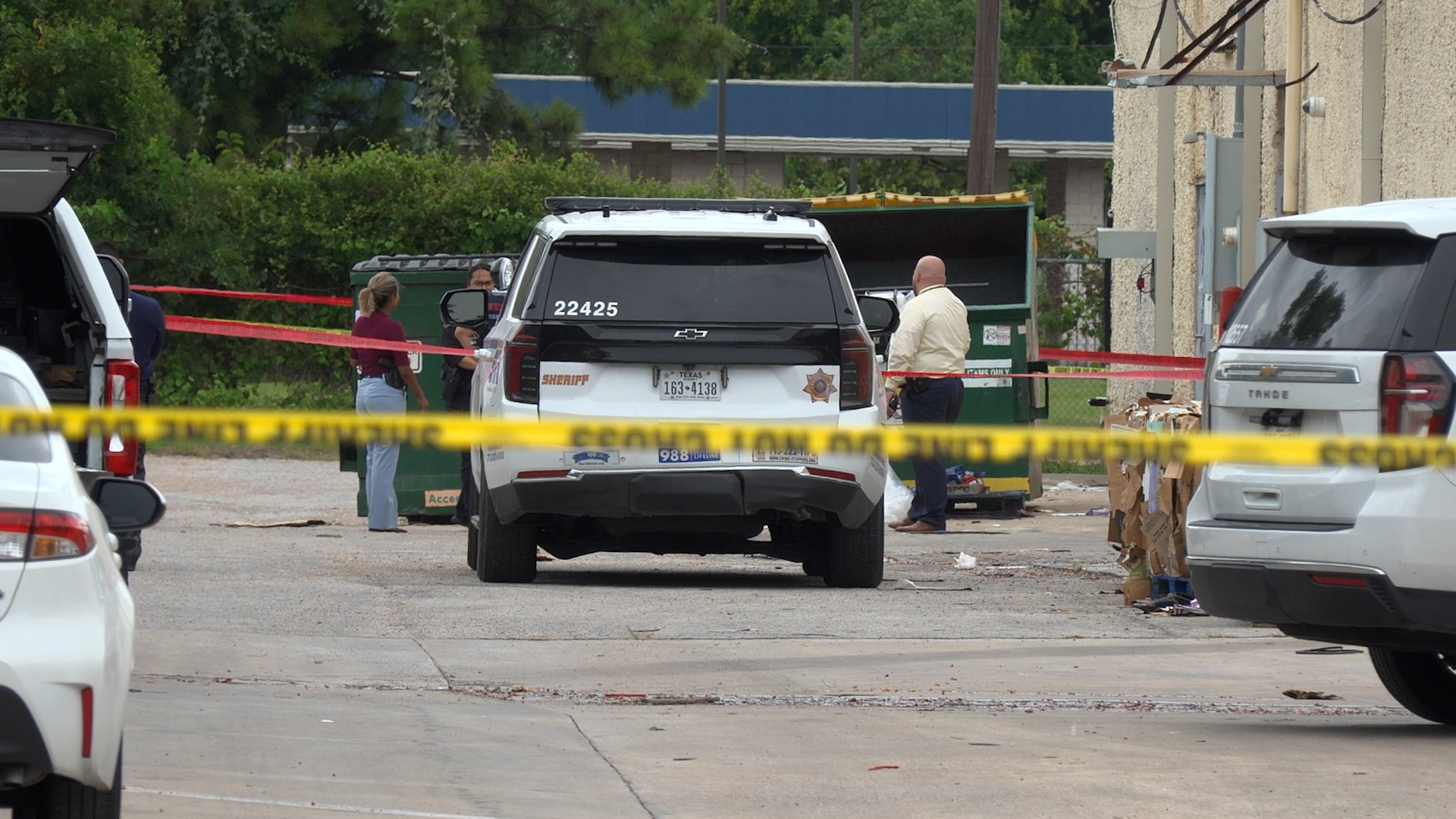 Sheriff SUV with number 22425 at a taped-off crime scene, investigators and bystanders in a lot with dumpsters.
