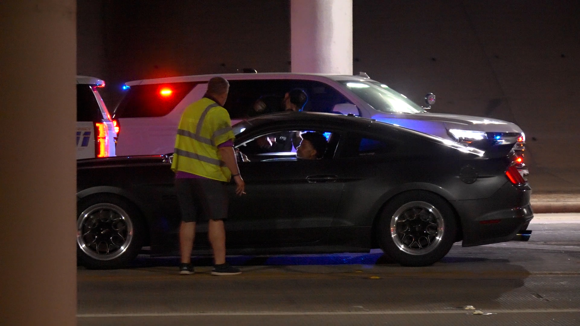 Nighttime traffic stop: a man in a high-visibility vest speaks to the driver of a dark car while police SUVs flash blue and red lights in the background.