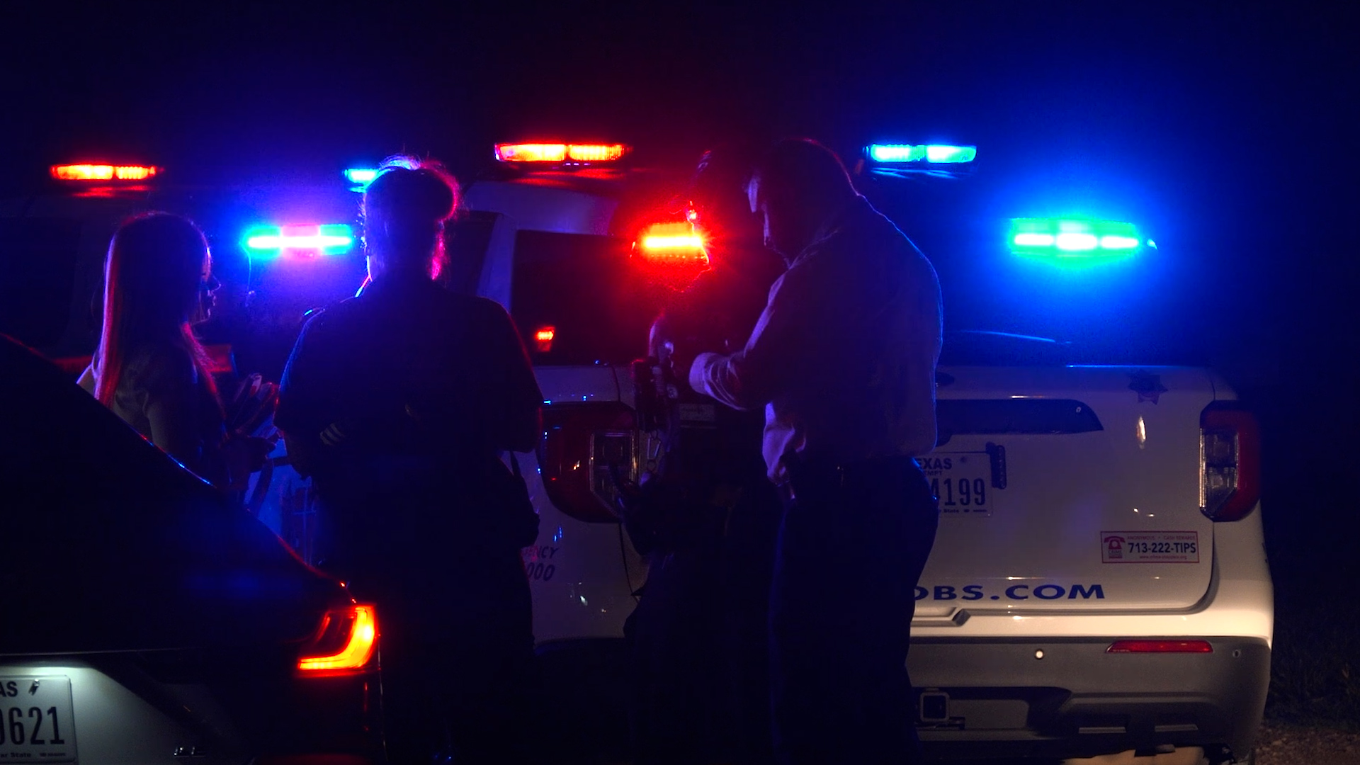 People stand beside a patrol SUV with red and blue emergency lights flashing at night.