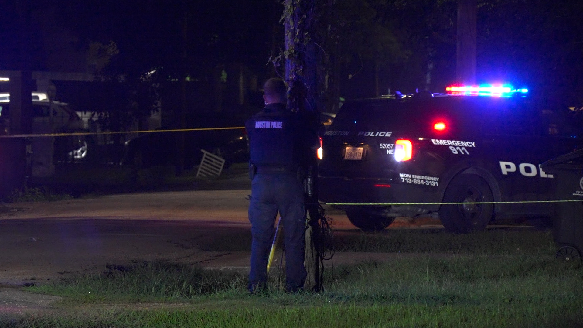 Houston Police SUV with flashing red and blue lights at night, officer facing away behind yellow caution tape on a cordoned street