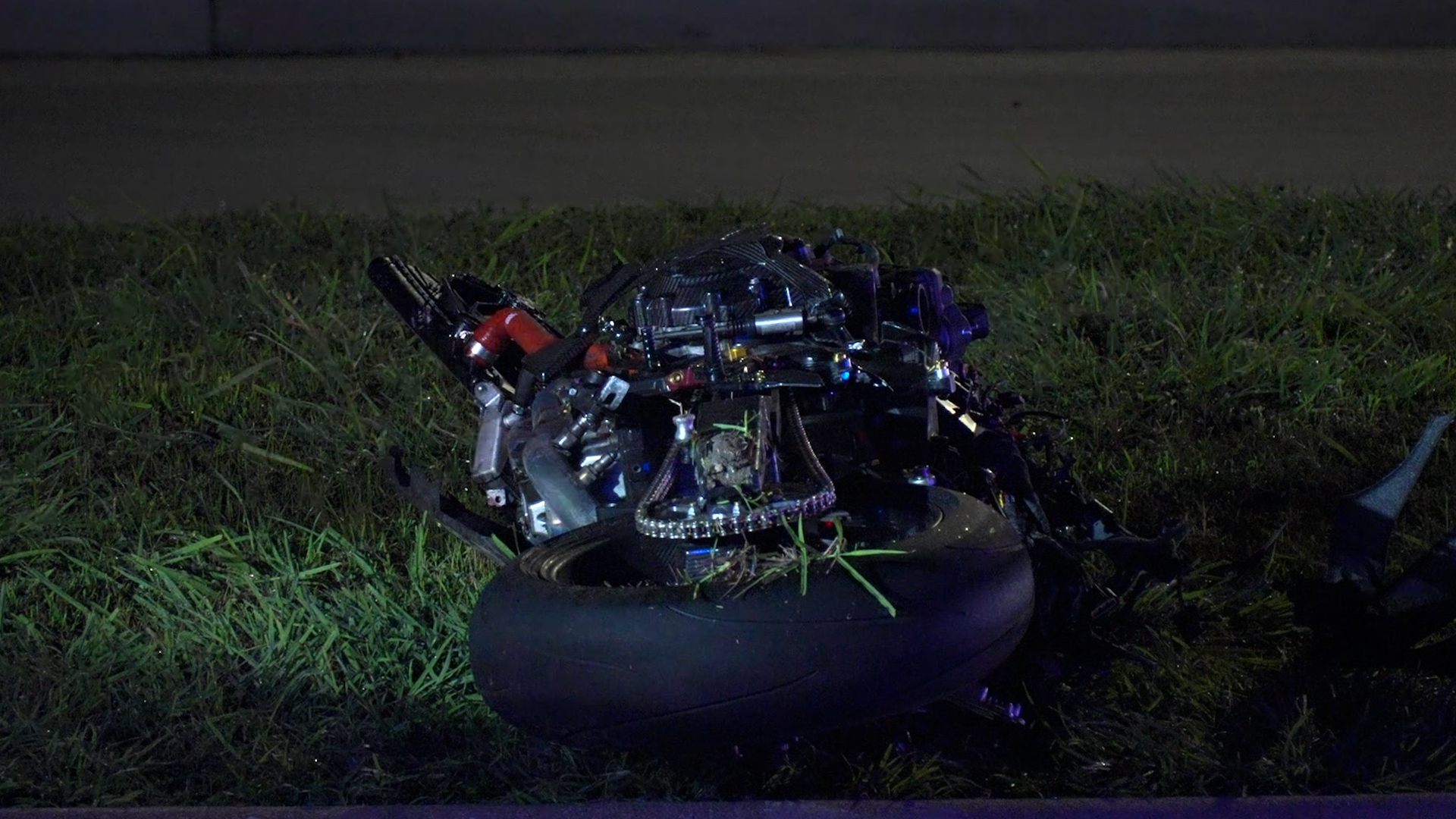 Nighttime roadside scene with a damaged motorcycle engine and wheel on a grassy verge.