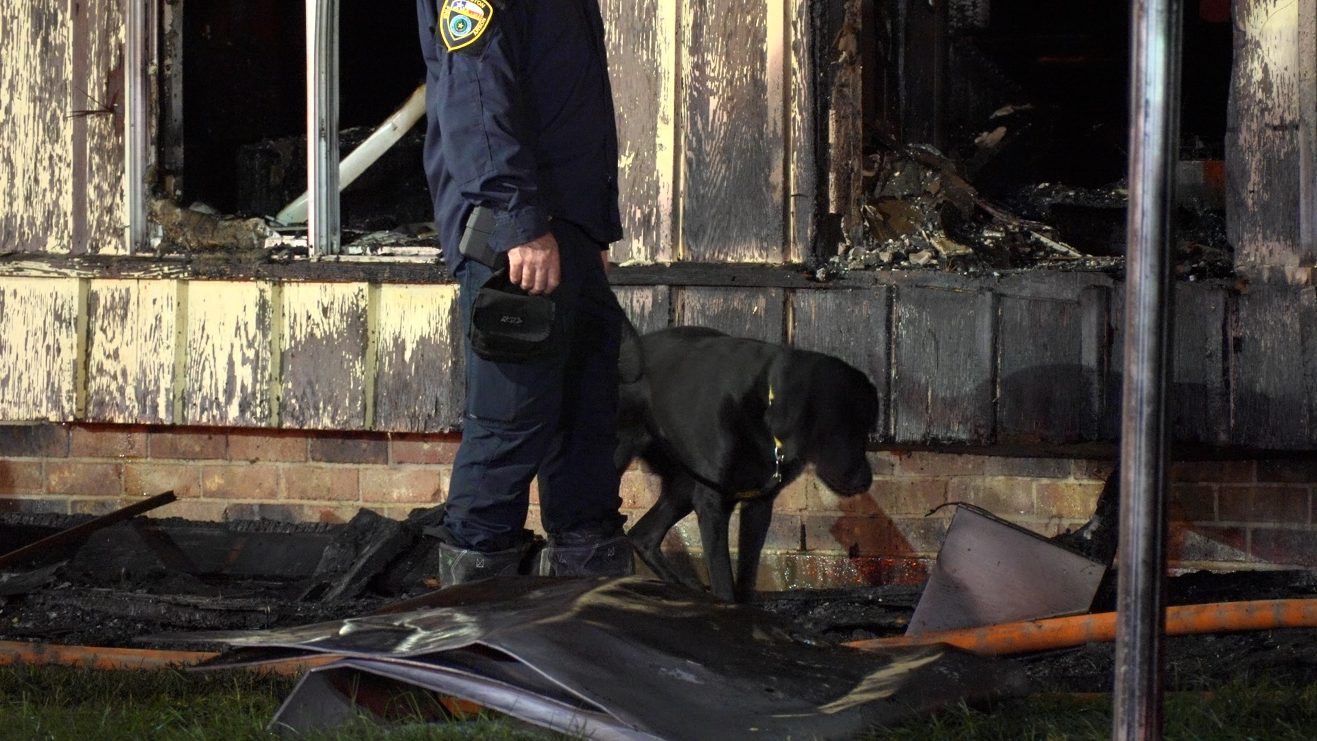 Firefighter in uniform with badge stands beside a black dog amid charred, burnt building debris.