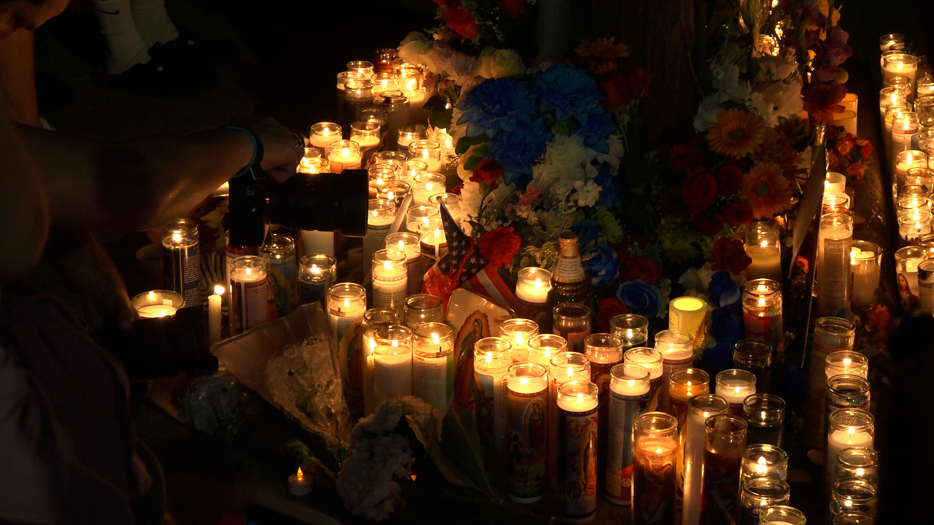 Memorial shrine with dozens of lit candles, colorful flowers, and small American flags in a dark setting.