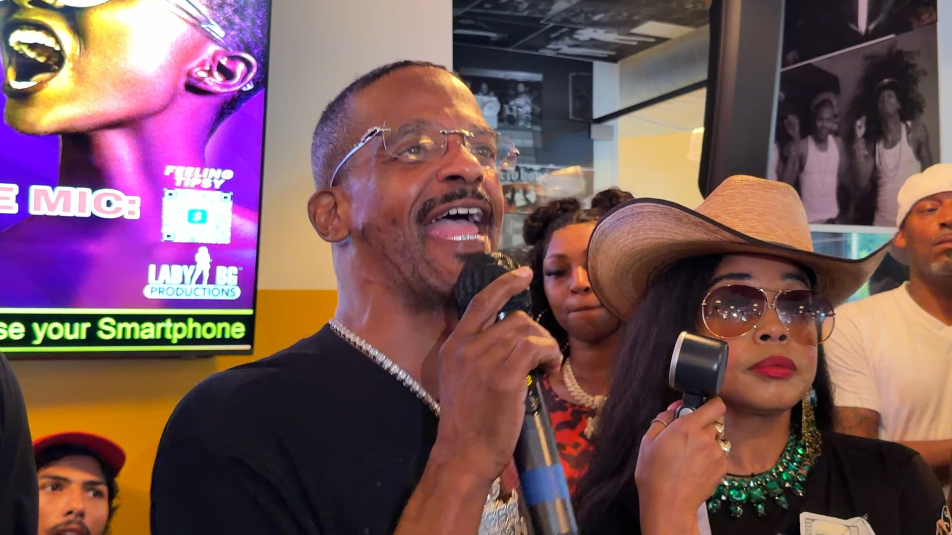 Man singing into a microphone at a colorful indoor event, with others in the background.