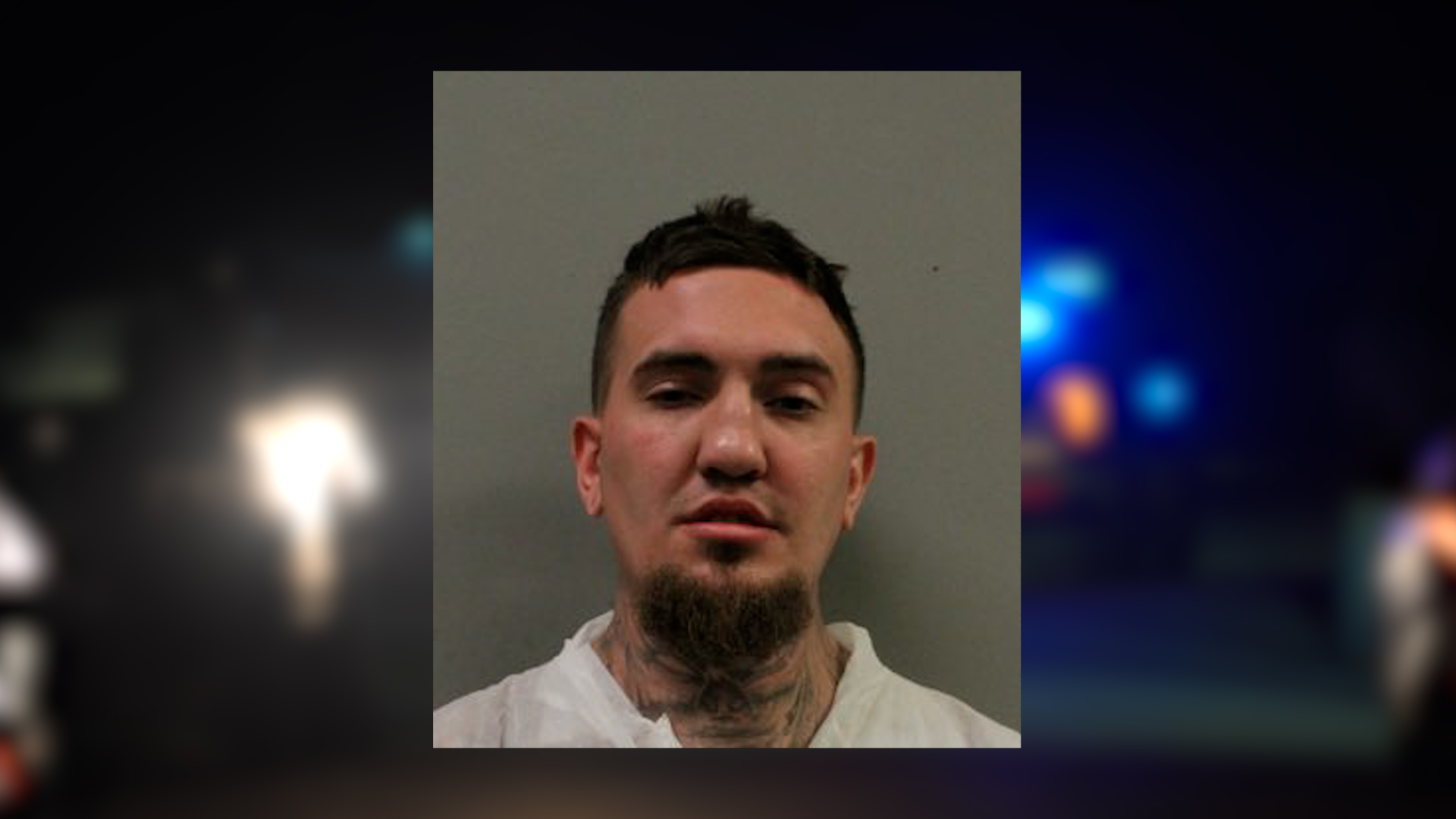 Mugshot-style photo of a man with a goatee and neck tattoo wearing a white shirt, standing against a plain gray background, with blurred police lights on the sides in the frame.