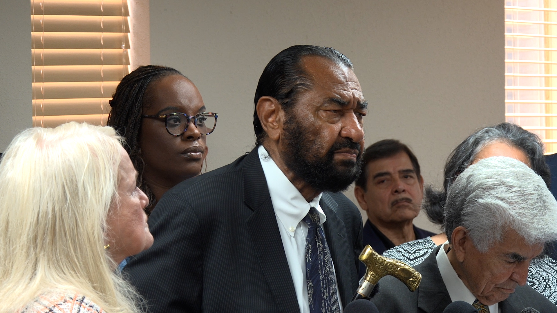 Man in dark suit and patterned tie speaks at a press event, surrounded by seated people in a room with blinds in the background.