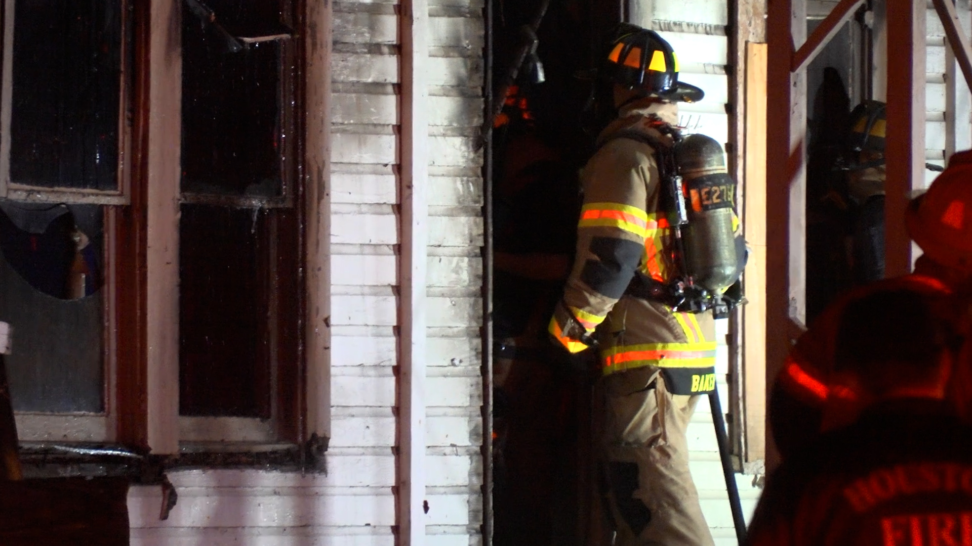 Firefighter in full turnout gear and air tank entering a smoke-filled doorway at night.