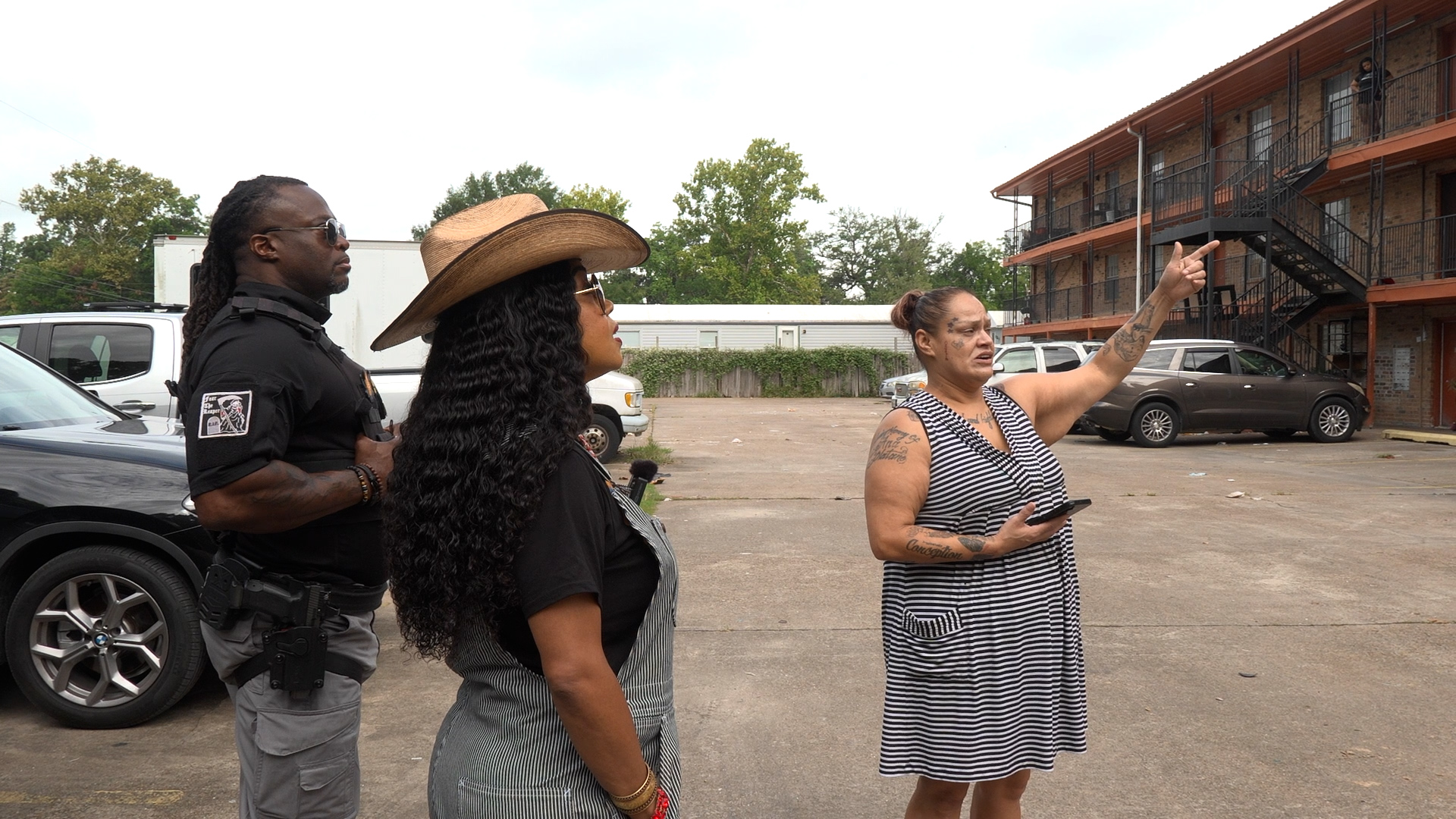 Three people stand in a parking lot near an apartment building; a police officer listens as two women converse, one gesturing with her arm raised.