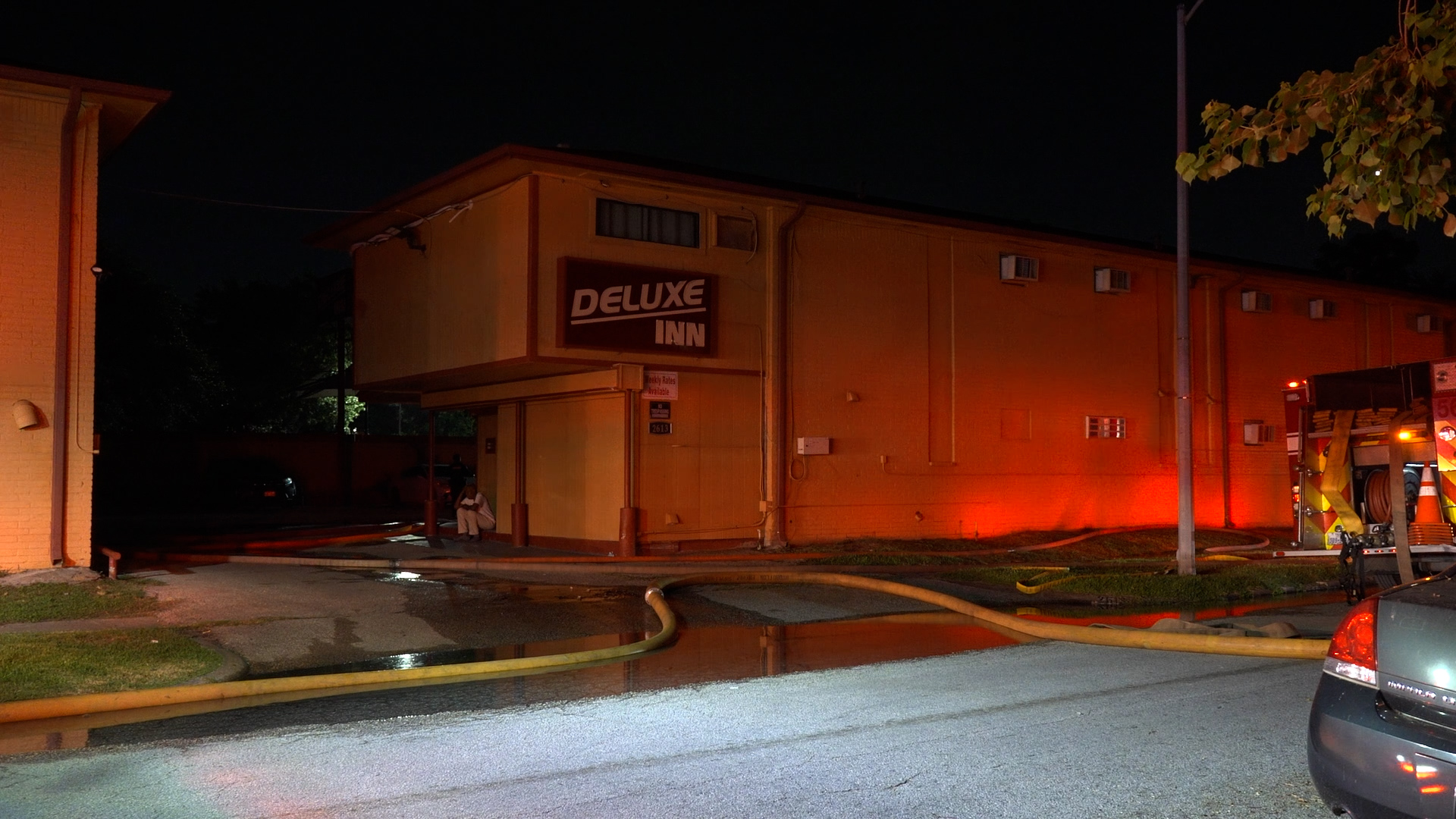 Night exterior of a motel with 'Deluxe Inn' sign, red emergency lighting, and coiled fire hoses on the wet pavement.