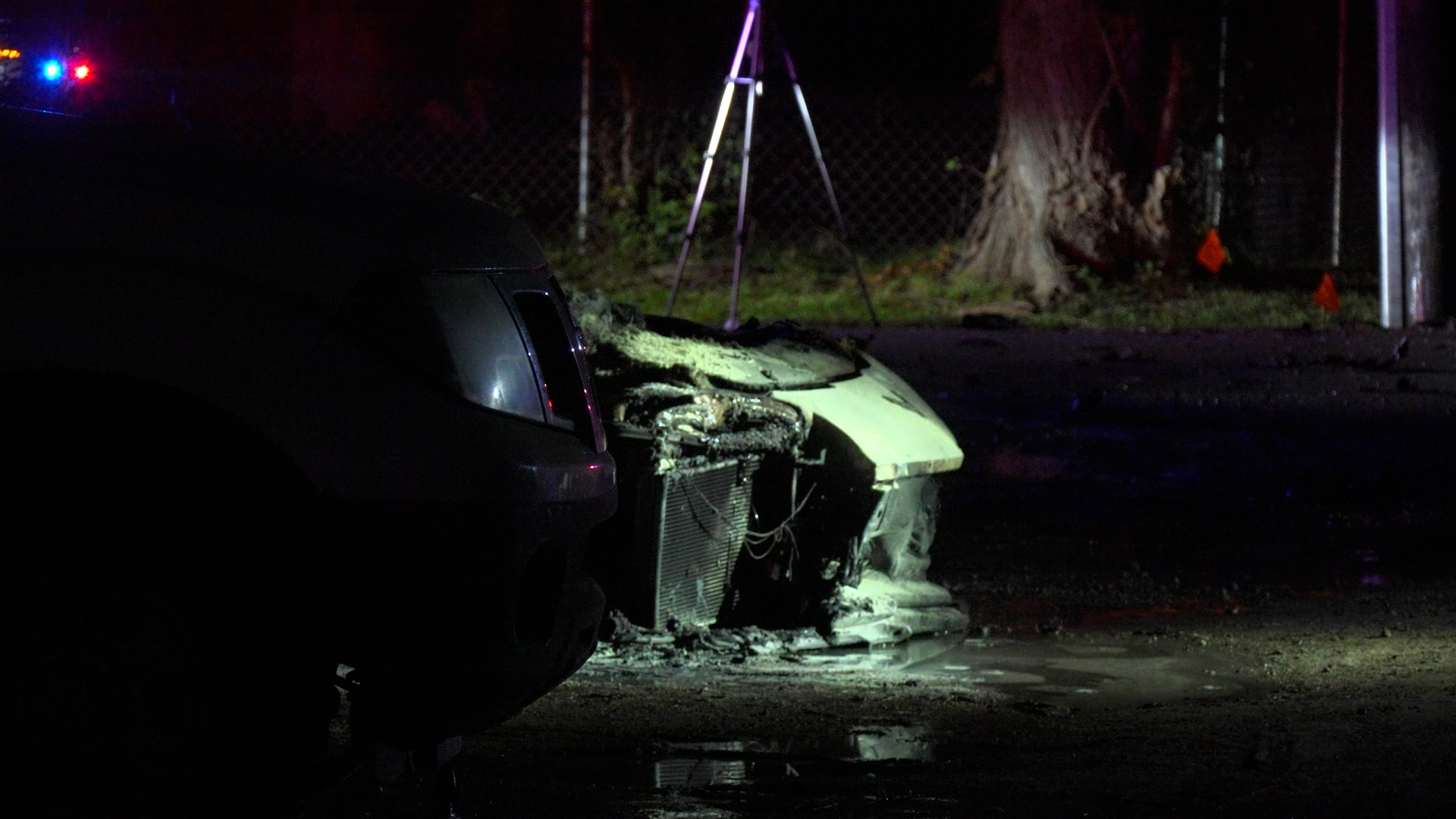 Nighttime crash scene with a heavily damaged car front-end, water or debris on the ground, and police lights in the distance behind a fence.