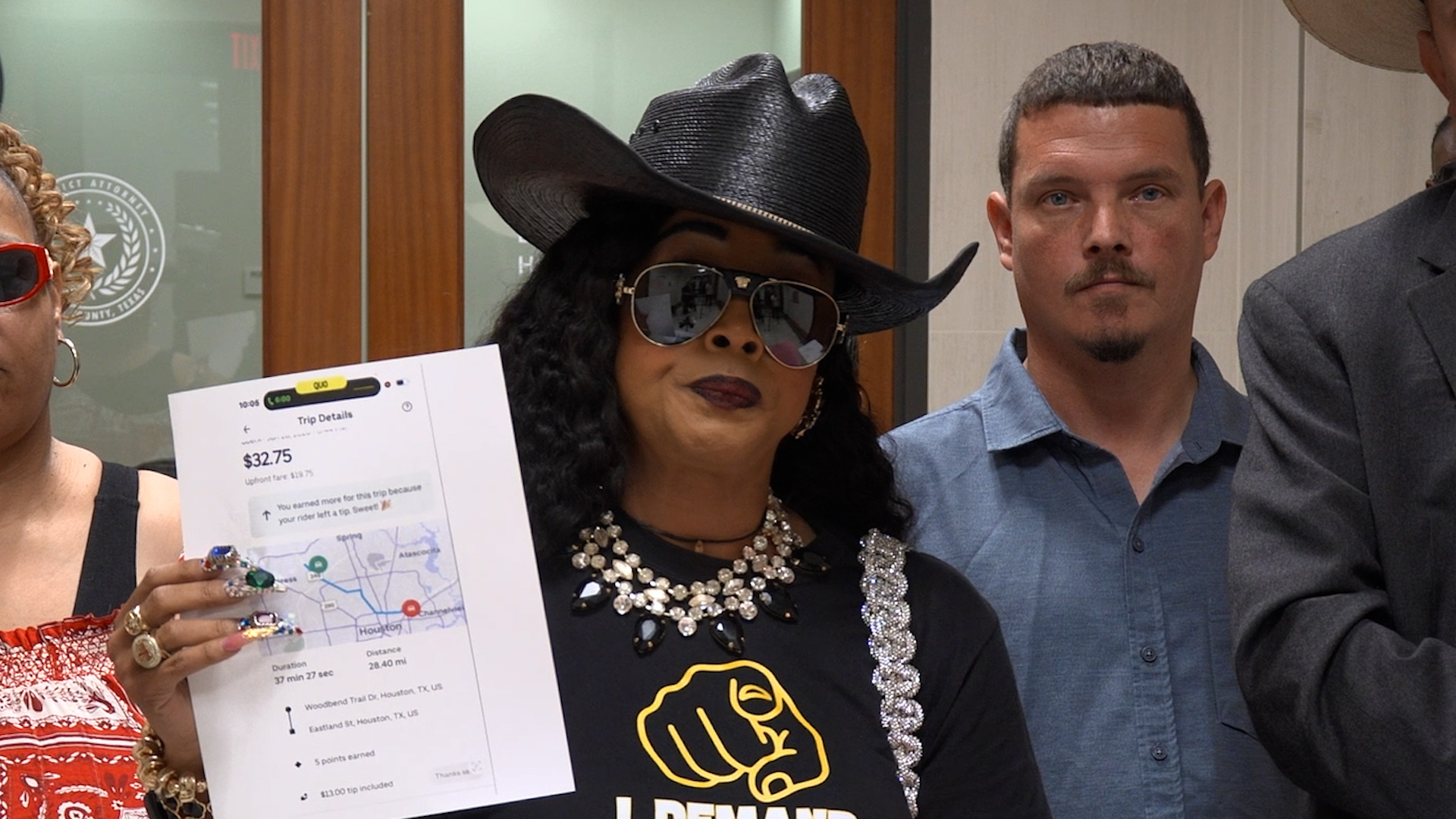 Group of people posing together; woman in a black hat and sunglasses holds a paper showing trip details and earnings, with a map visible on the page.