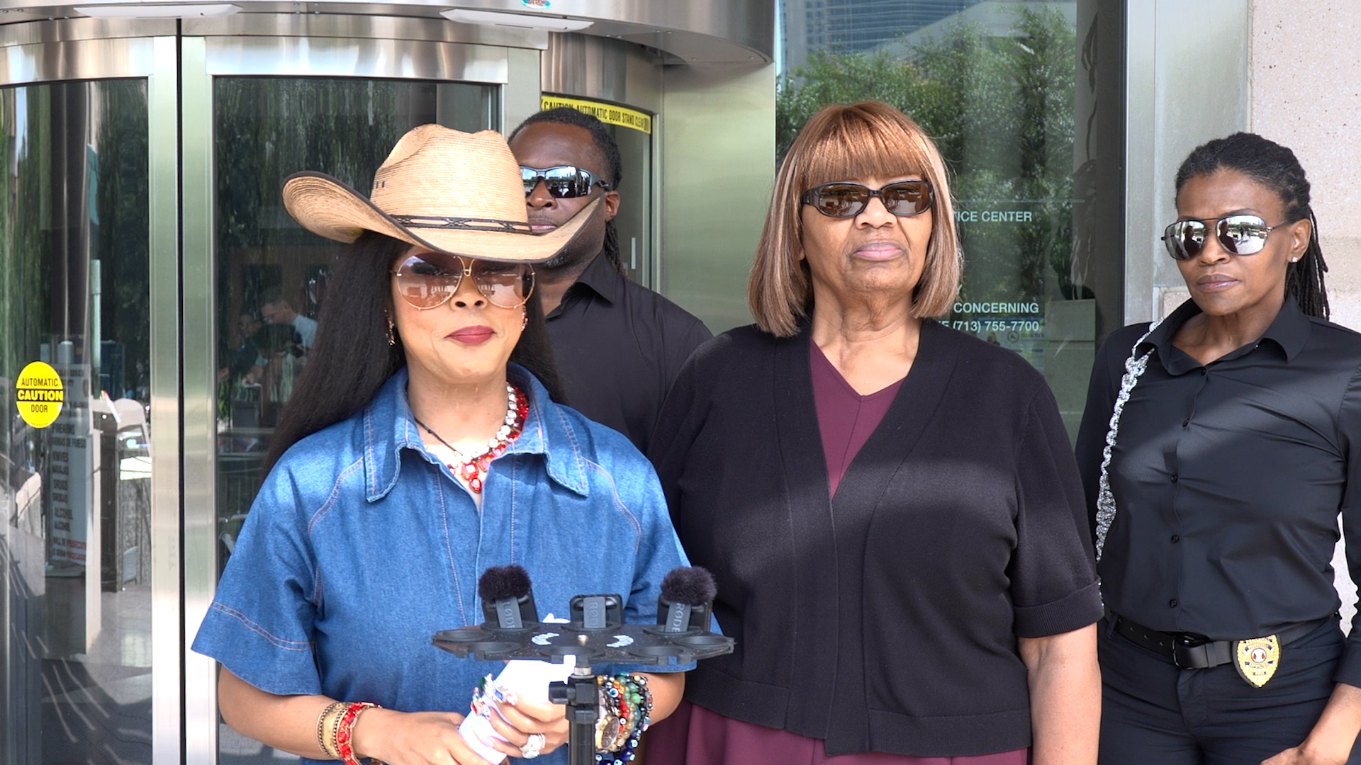 Group of four adults posing outside a glass-front building; woman in a denim shirt wearing a wide-brim hat and sunglasses stands at left with microphones in front.