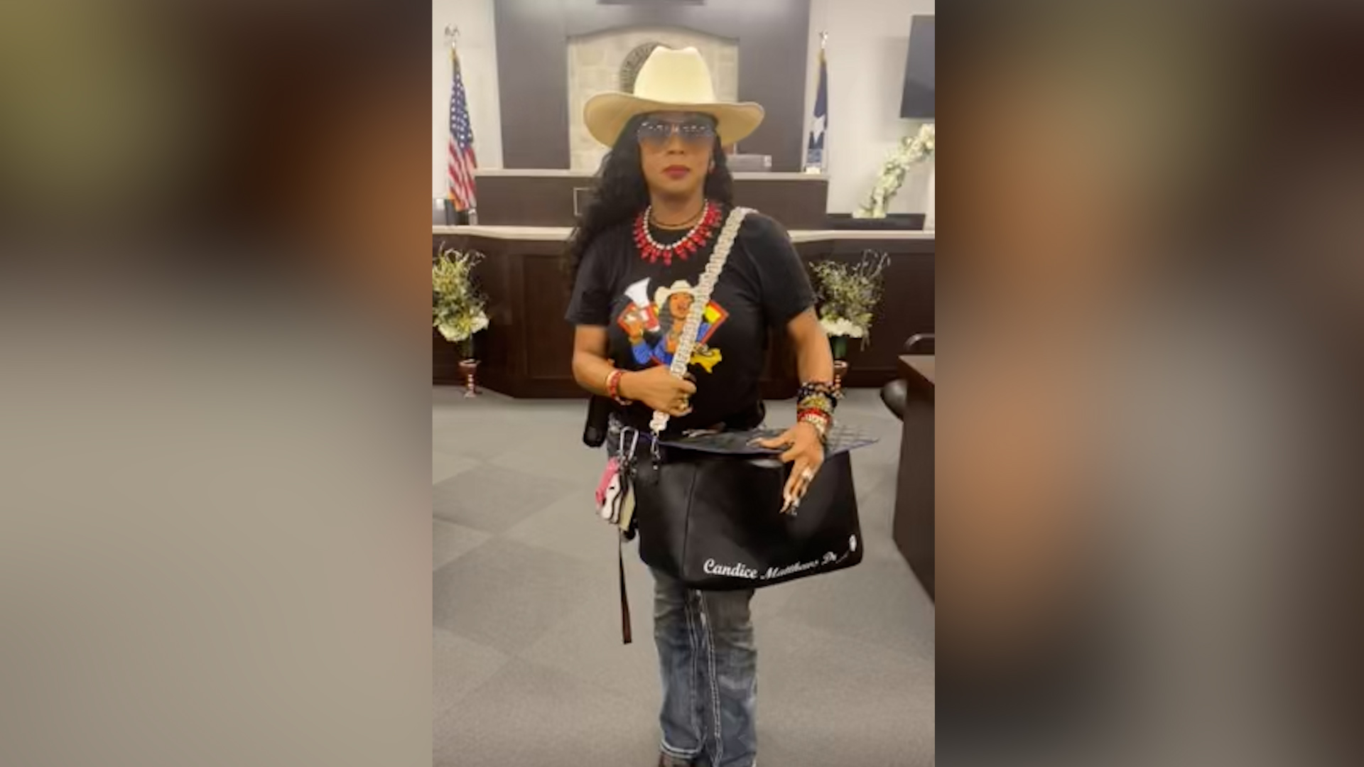 Person wearing a tan cowboy hat, sunglasses, red beaded necklace, and black shirt with a colorful graphic, standing in a lobby while holding a white strap and a black tote bag with white text.
