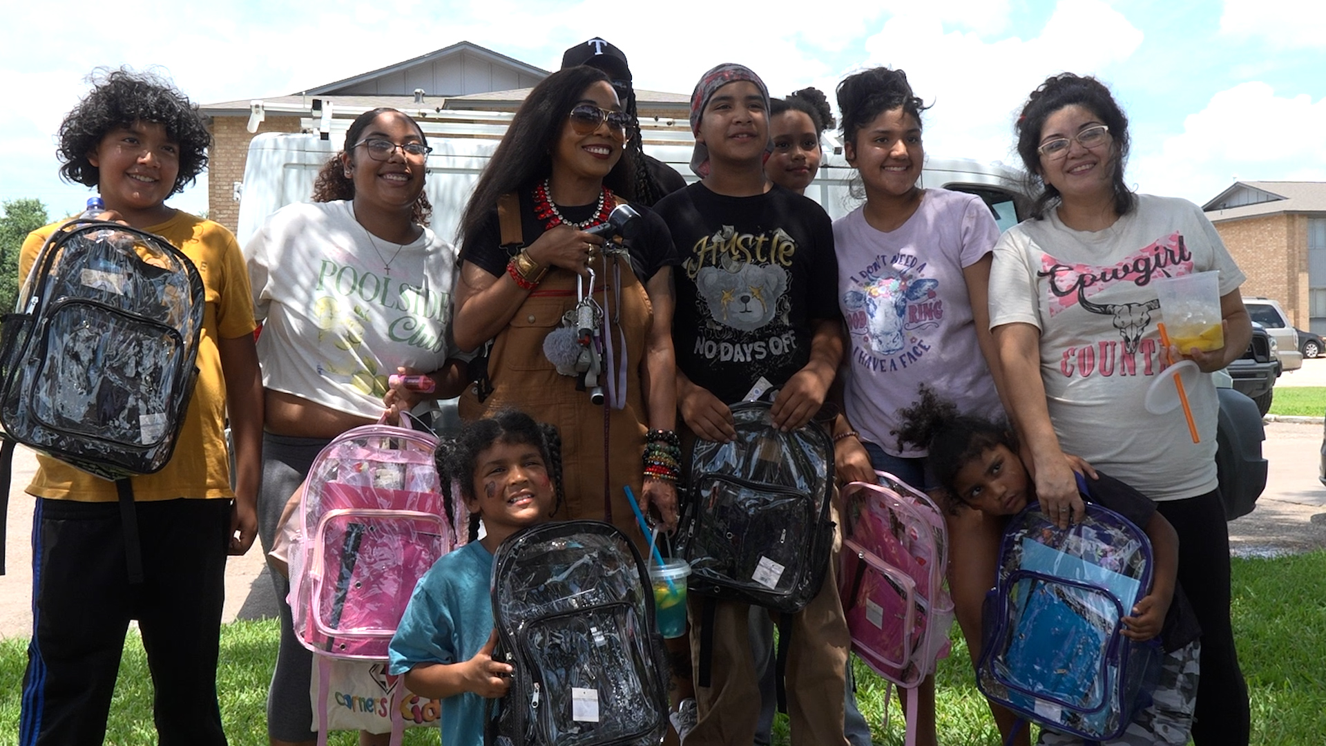Smiling group of diverse children and a woman posing outdoors with colorful backpacks in a residential area.