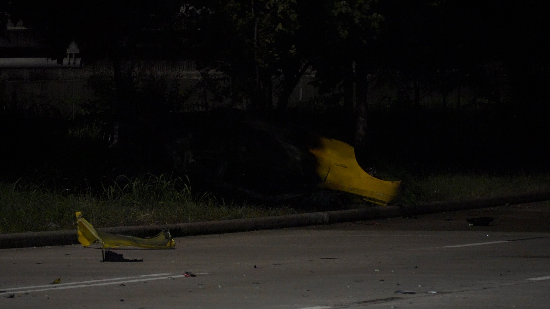 Nighttime street scene with torn yellow caution tape and a damaged yellow barrier along the curb; grass and trees in the background.