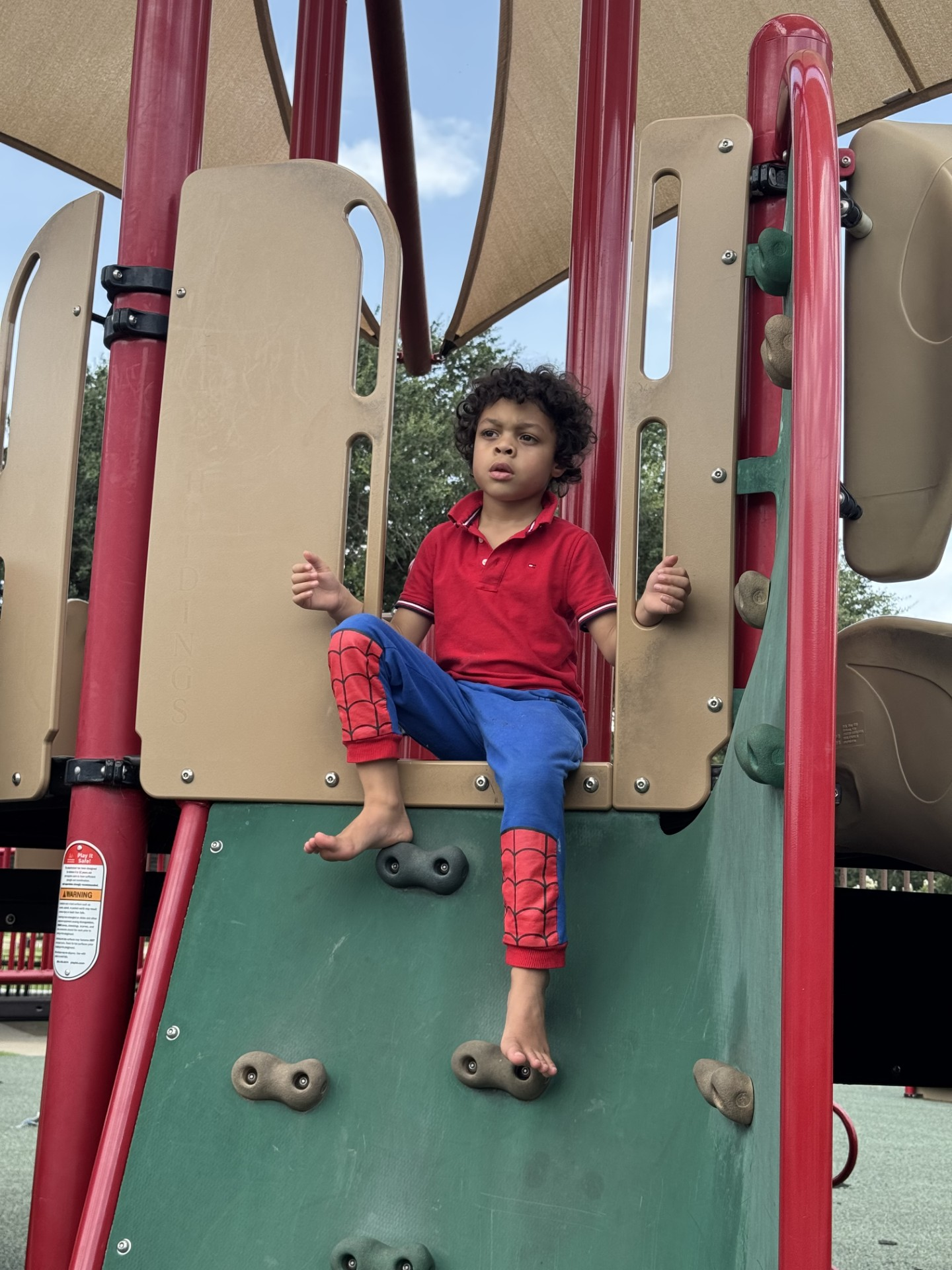 Young child in a red Spider-Man outfit sits on a playground climbing wall.