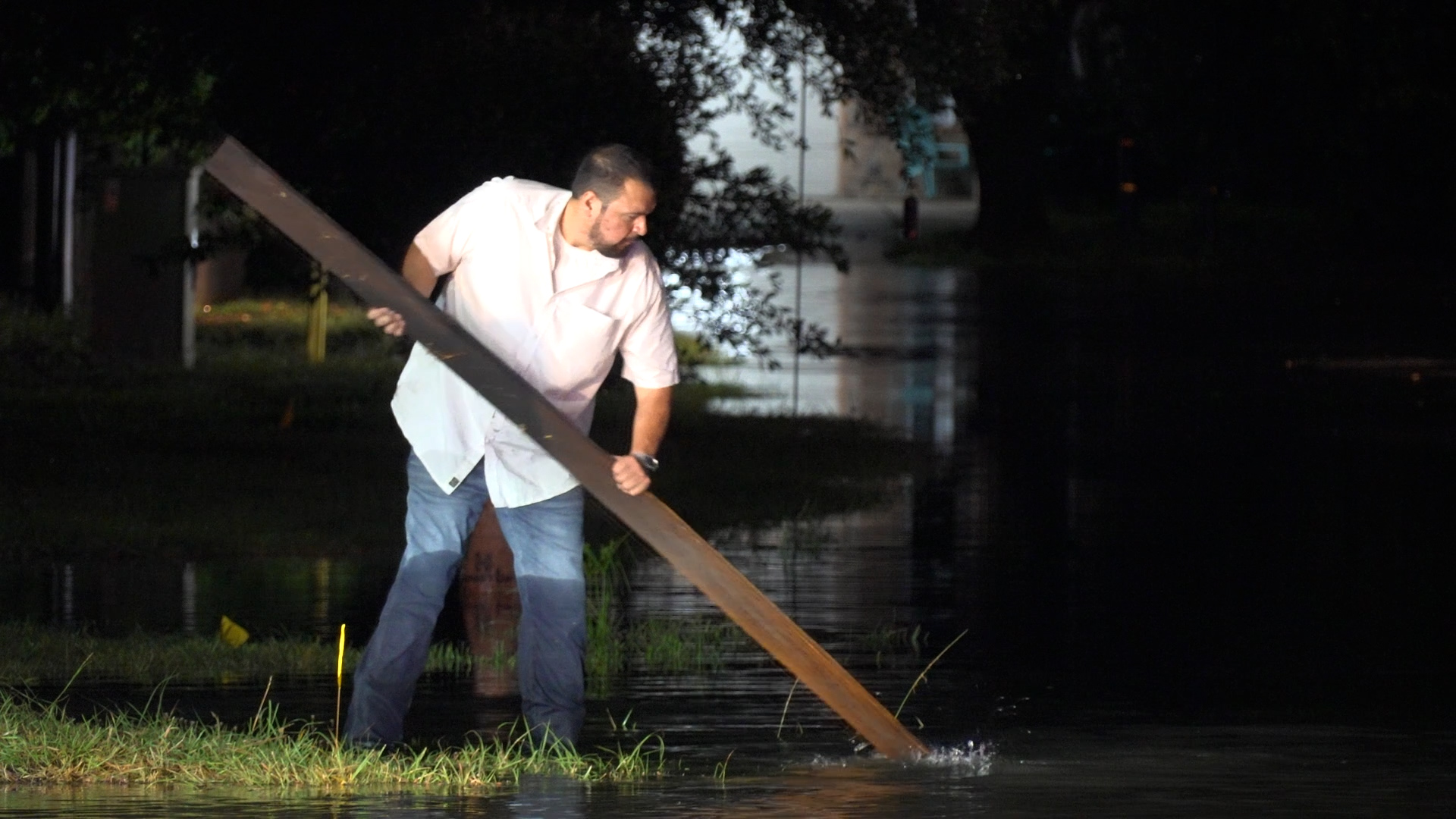 Man in a white shirt and jeans stands in shallow water at night, holding a long wooden plank.