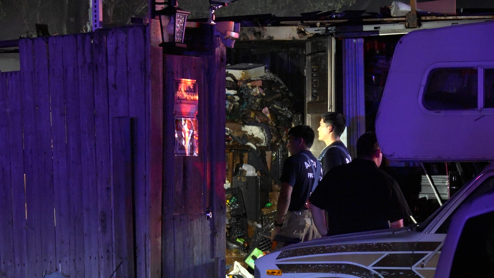 Nighttime scene with firefighters by an open trailer filled with trash and debris, illuminated by purple emergency lights.