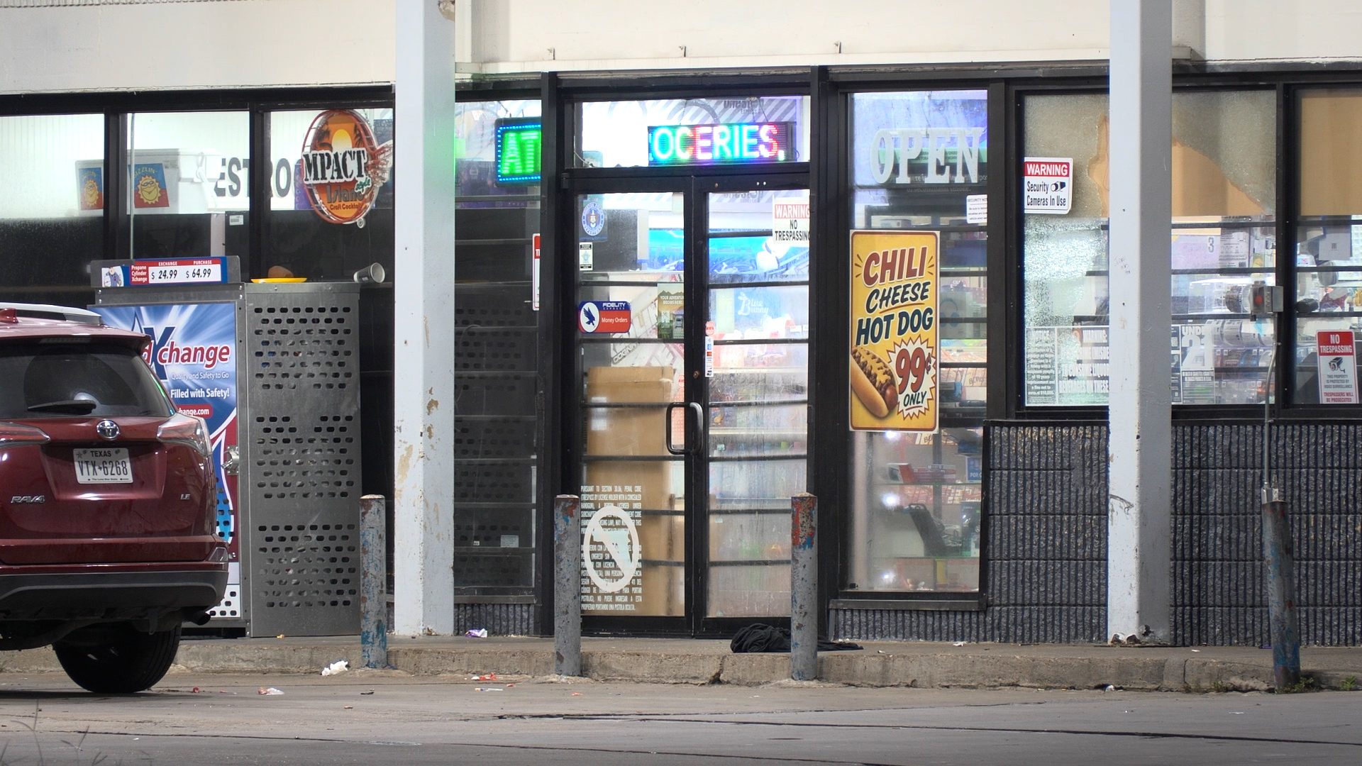 Front of a convenience store with a neon OPEN sign and a bright chili cheese hot dog poster in the window, car parked outside.