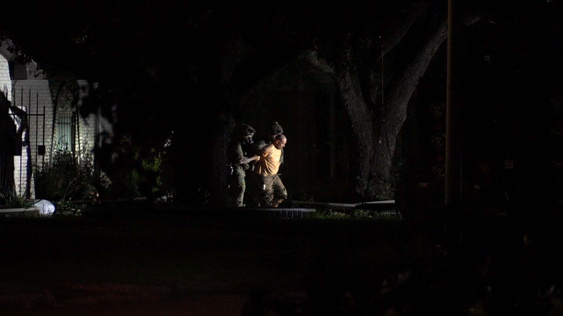 Night scene on a residential street: several people, including two officers, hold and escort a man in an orange shirt at night.