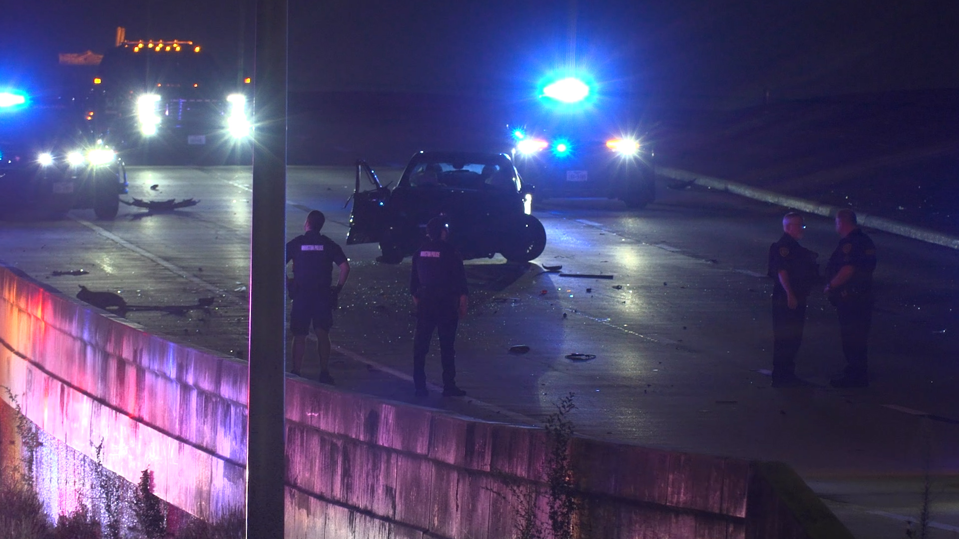 Nighttime highway crash scene with a damaged car, police officers, and flashing emergency lights behind a barrier.