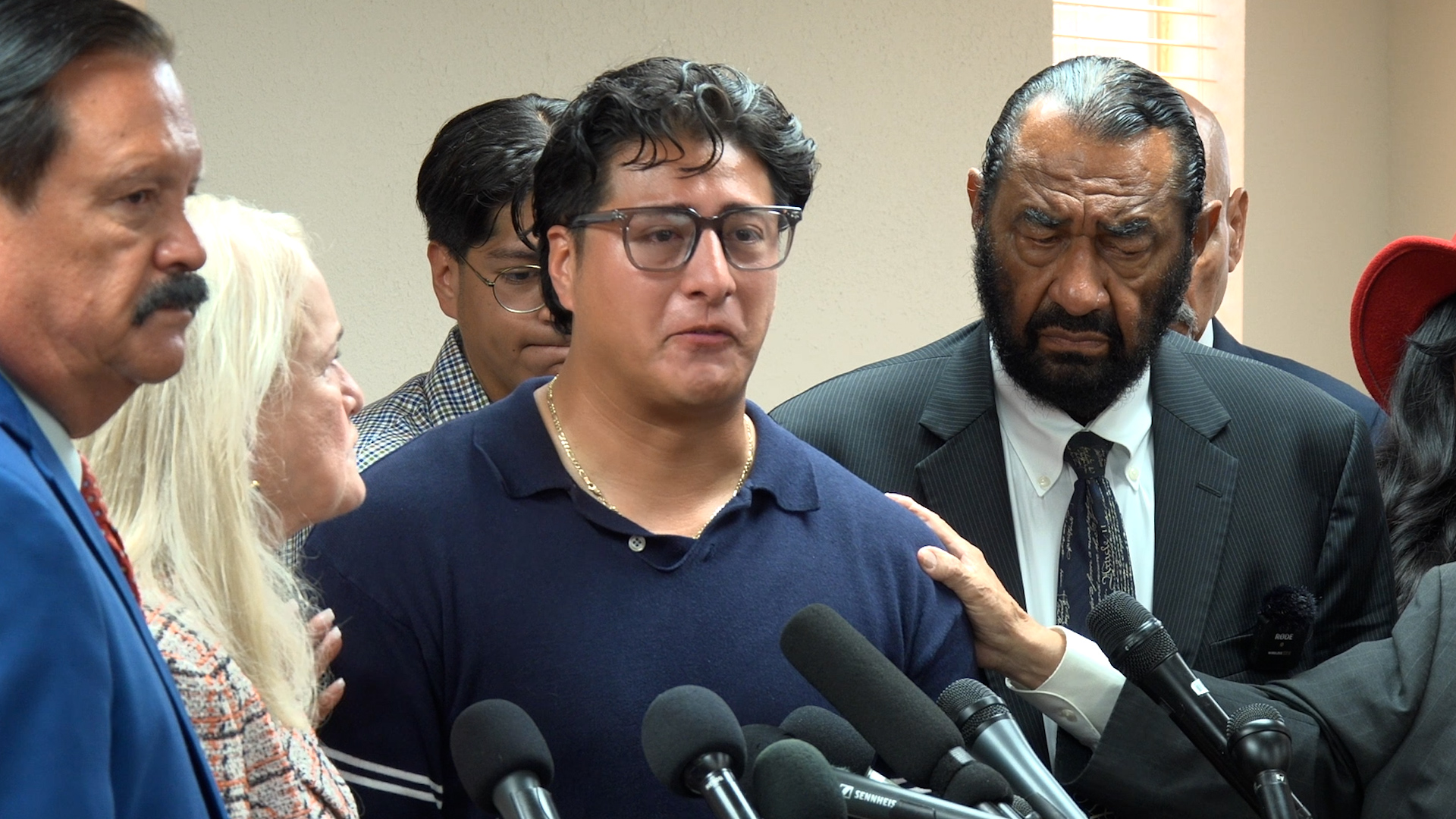 Man in a navy polo shirt speaks to reporters at a press conference, surrounded by multiple microphones and attentive bystanders.