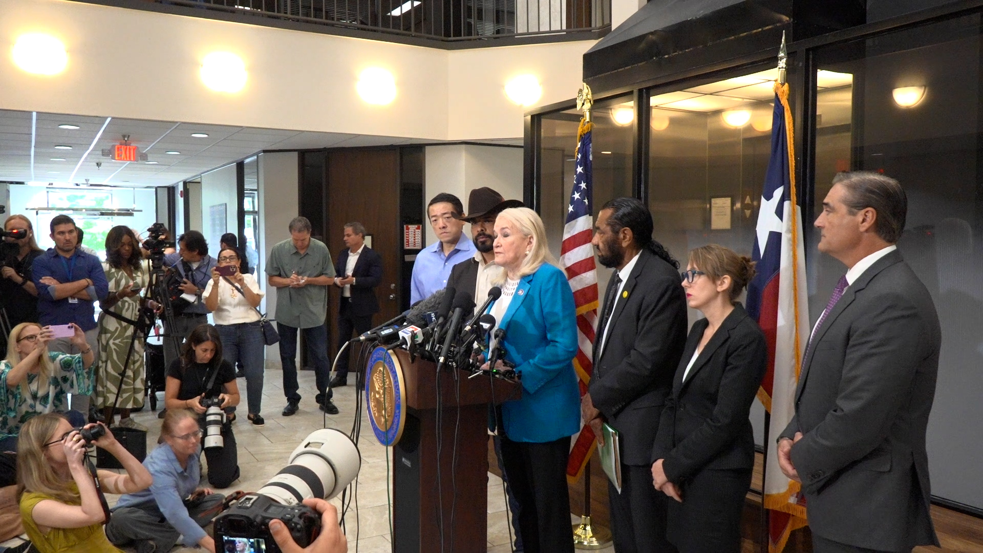 Female official in a teal blazer speaks at a podium with microphones during a press conference, surrounded by reporters and cameras.