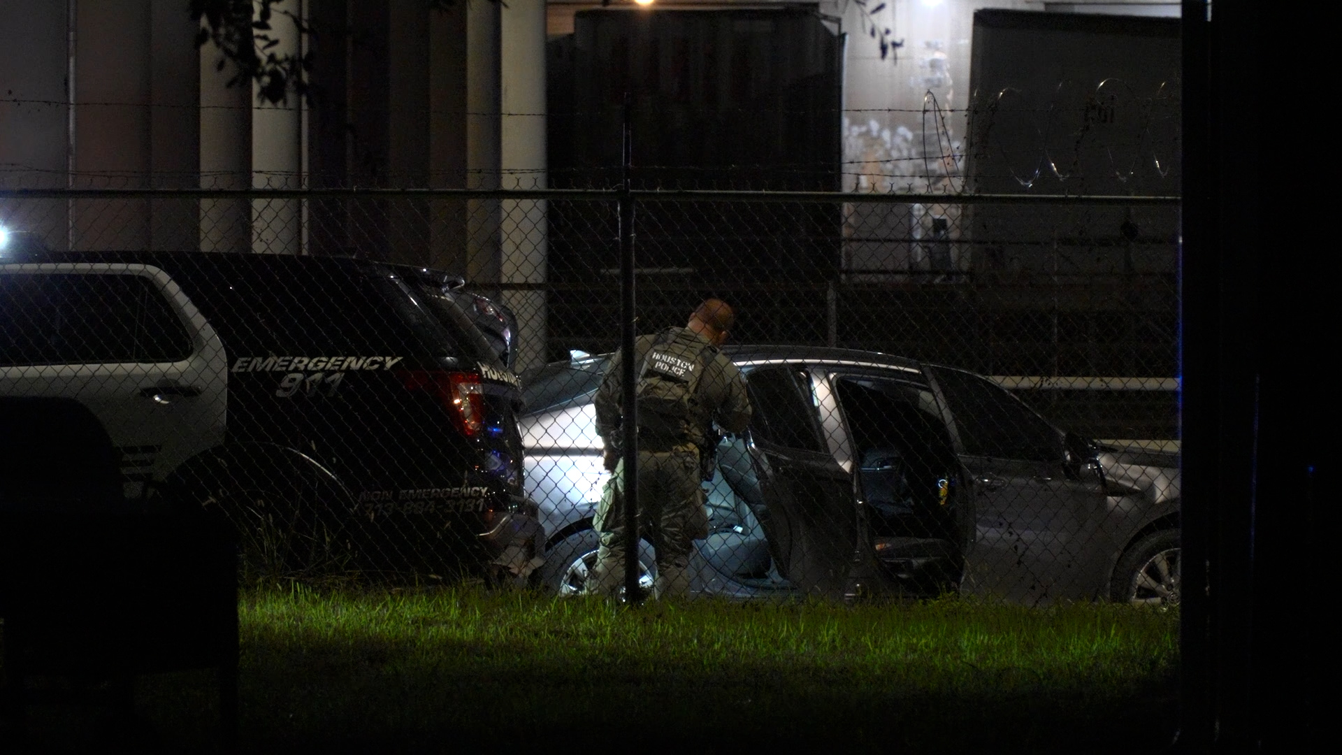 Night scene: a uniformed police officer stands by a car with its door open in a fenced, dimly lit yard; barbed wire fence in foreground and another emergency vehicle present nearby.