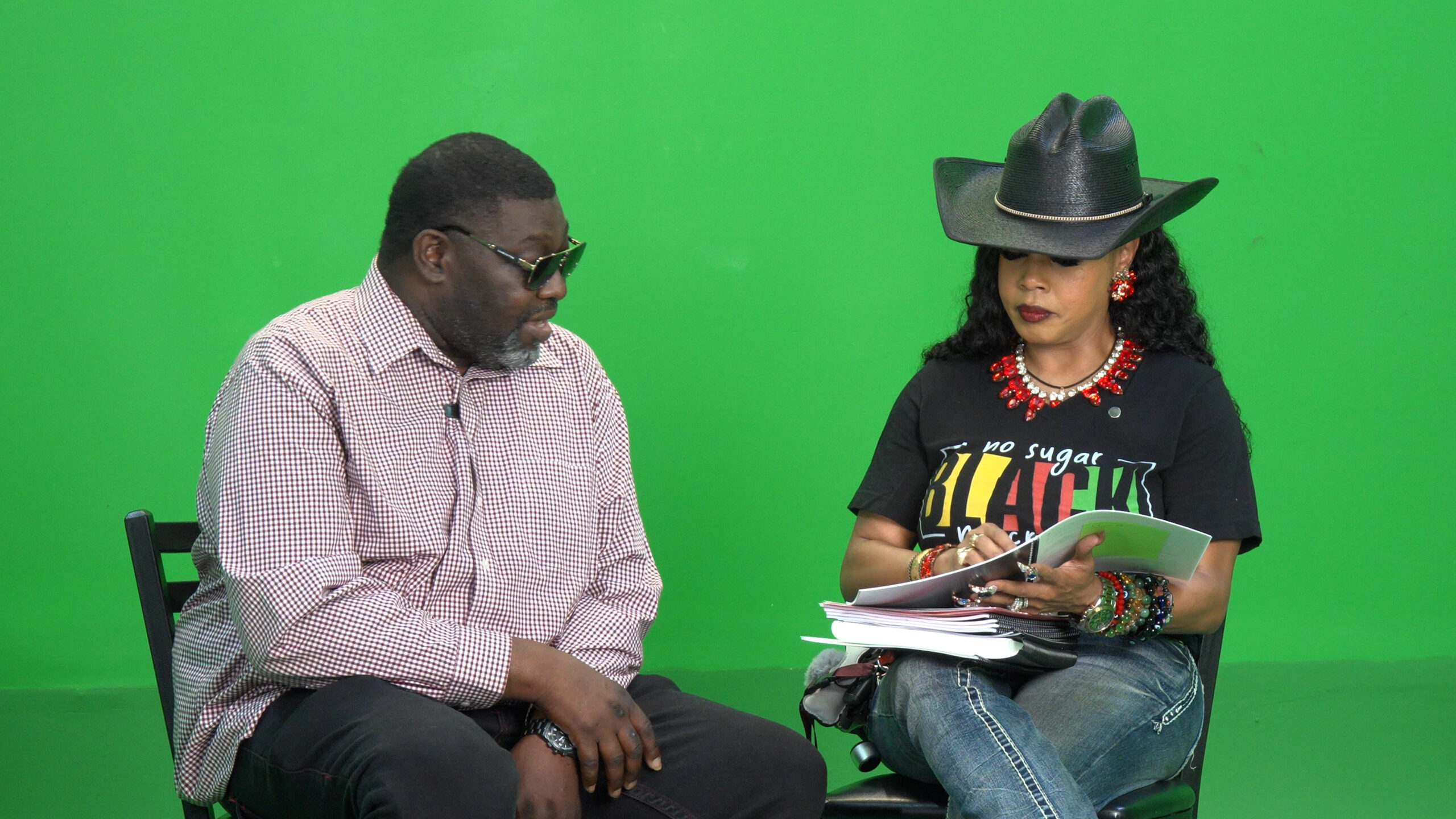 Two people sit side by side in a studio with a green screen; man in a checkered shirt and glasses talks while a woman in a large black hat holds papers.