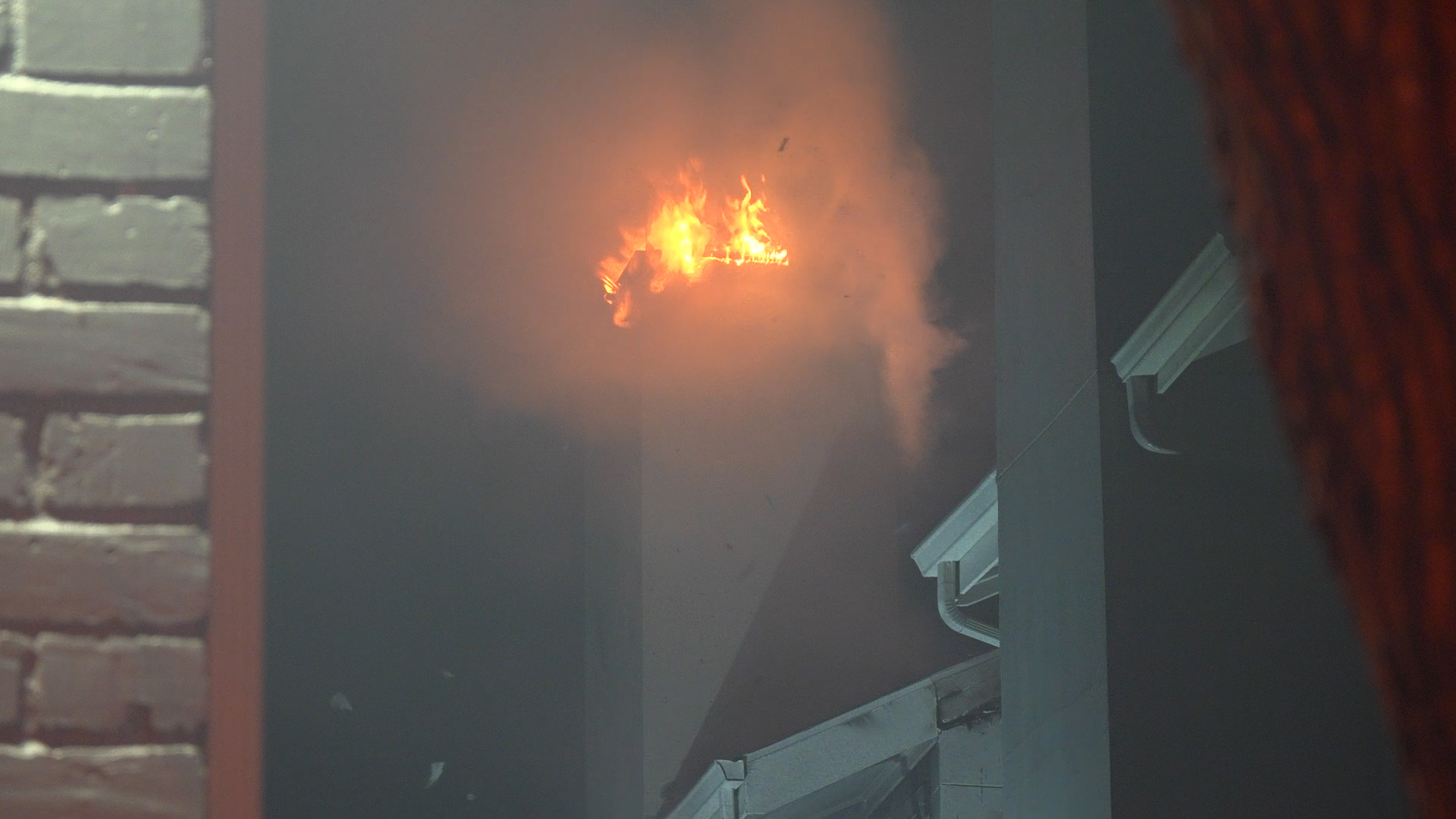 Flames and smoke billow from a doorway in a building, with a brick wall visible on the left and structural siding on the right.