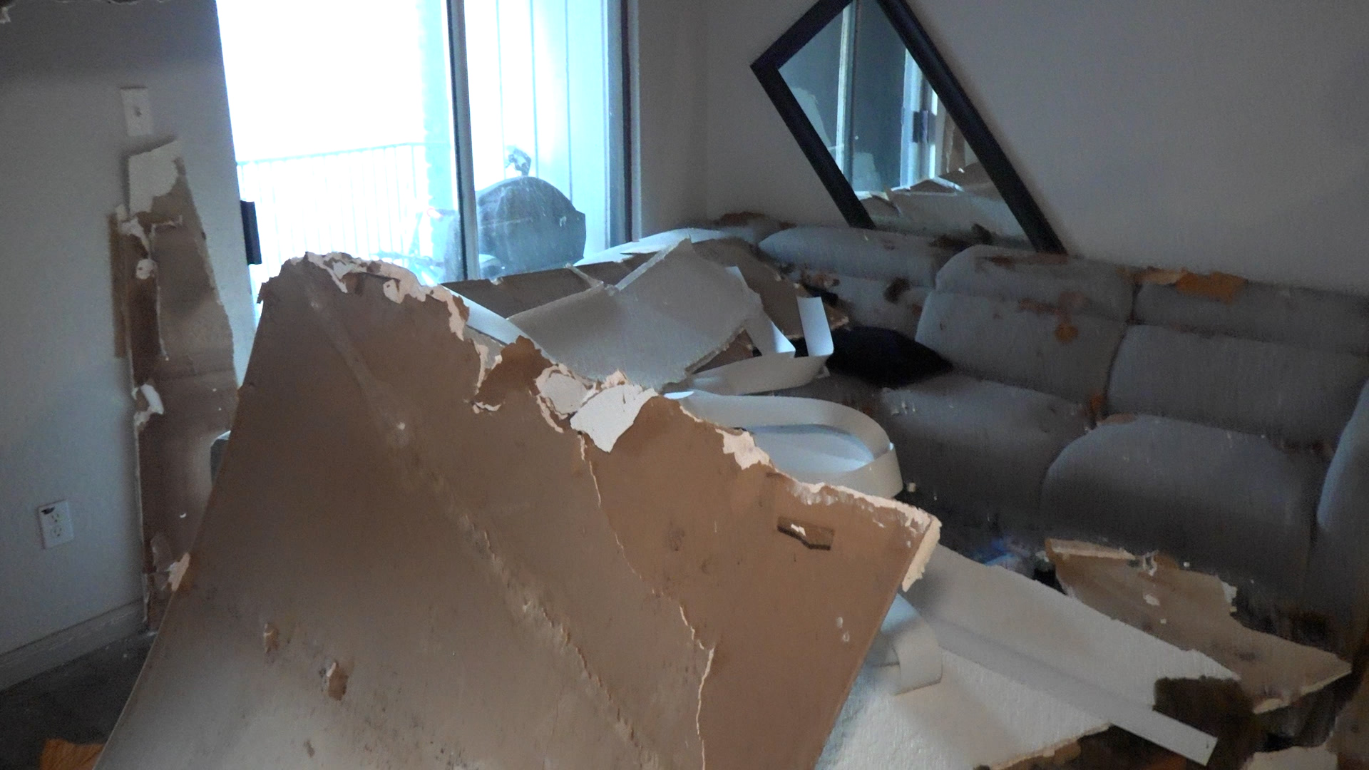 A badly damaged living room with collapsed drywall and debris piled on a couch and floor, near a large window showing a balcony outside.