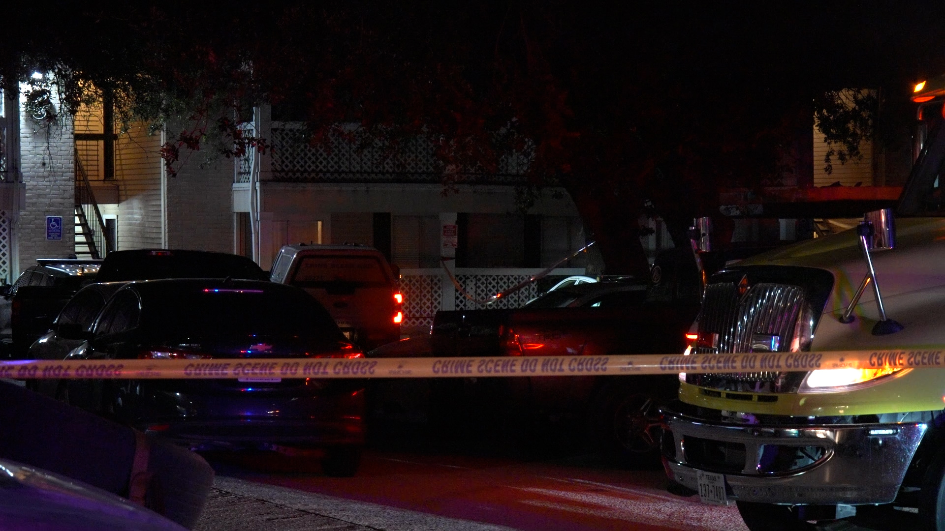 Nighttime street scene with crime-scene tape across the foreground and parked cars, emergency lights reflecting off wet pavement at a residential area night.