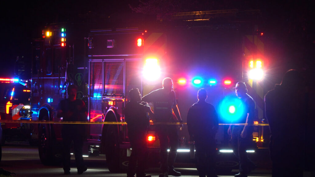 Emergency responders near a fire engine at night with flashing red and blue lights and police tape in the foreground.