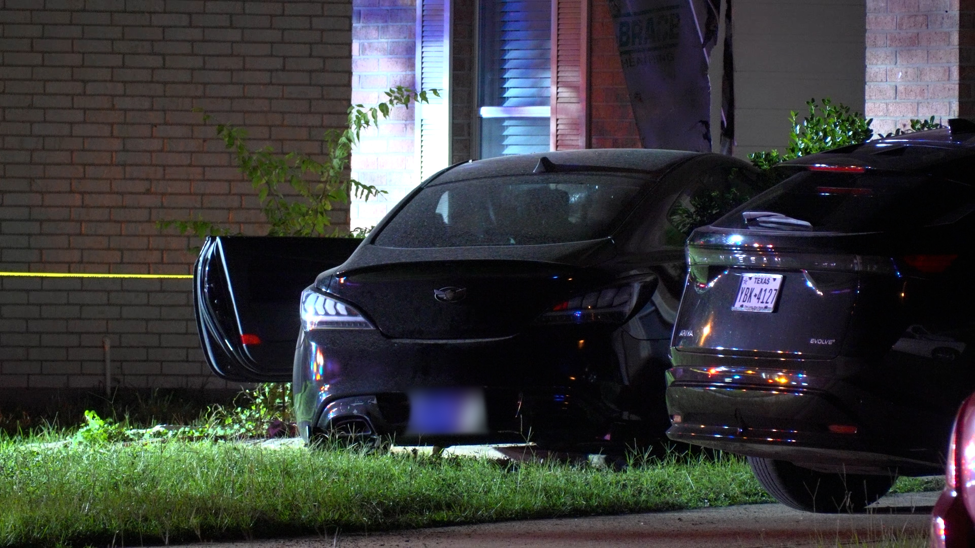 Nighttime crash scene with a black car and an open door, damaged front end, parked near a brick building; yellow caution tape in the background.