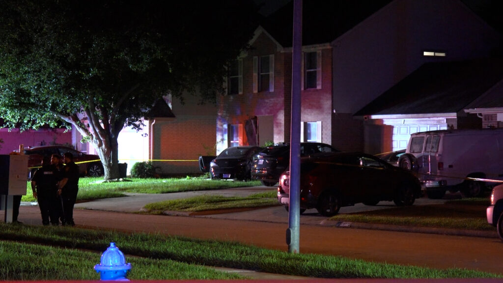 Nighttime residential street with yellow police tape, two officers, and several parked cars near a brick house under colorful lights on a crime scene.