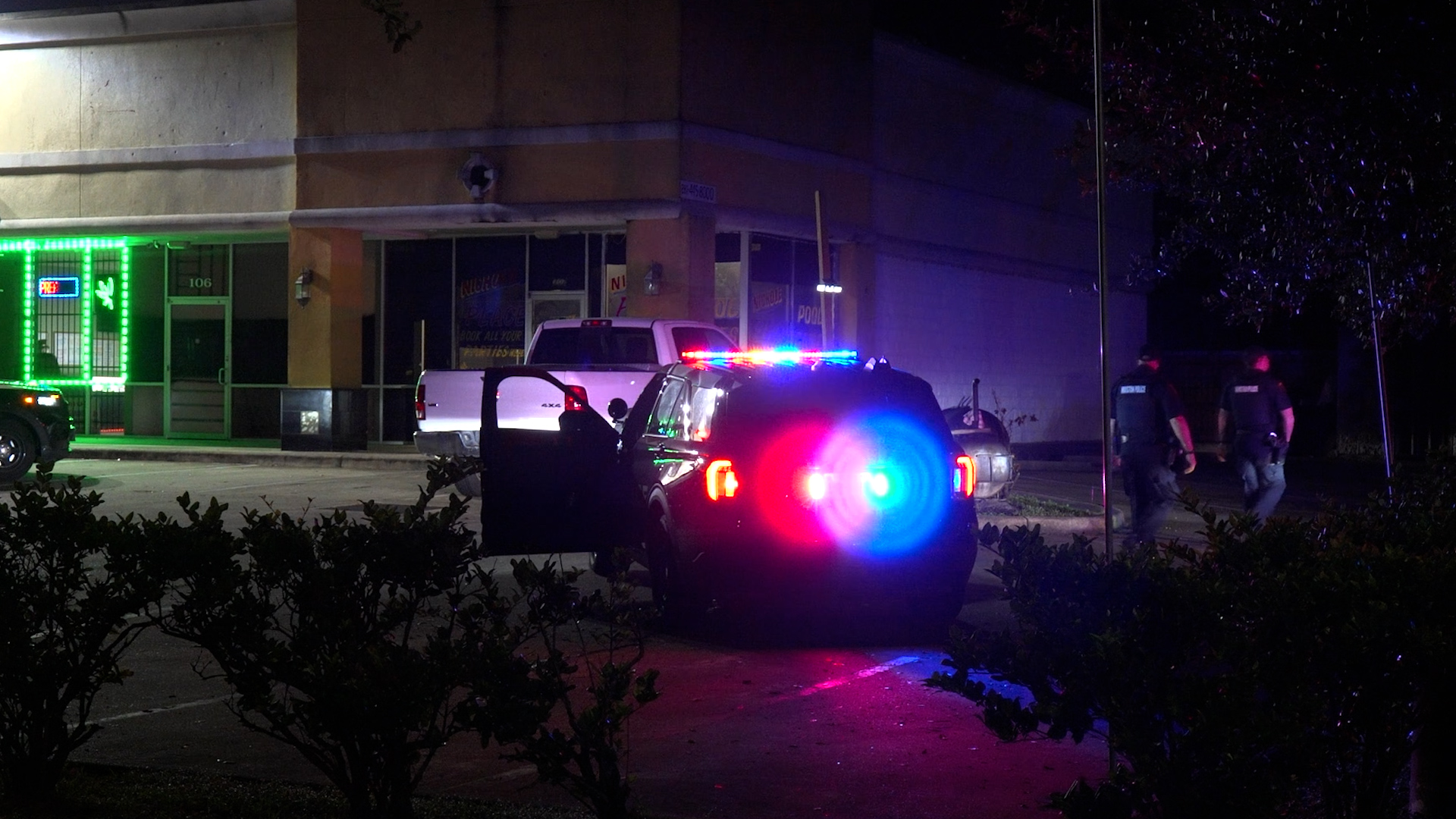 Police SUV with red and blue emergency lights flashing on a dark street at night, with storefronts in the background and a white pickup nearby.