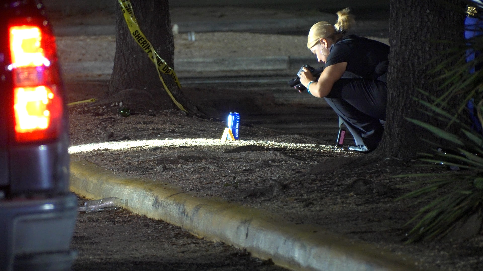 Photographer kneels at night to document a crime scene with yellow caution tape and a blue Bud Light can on the ground nearby.