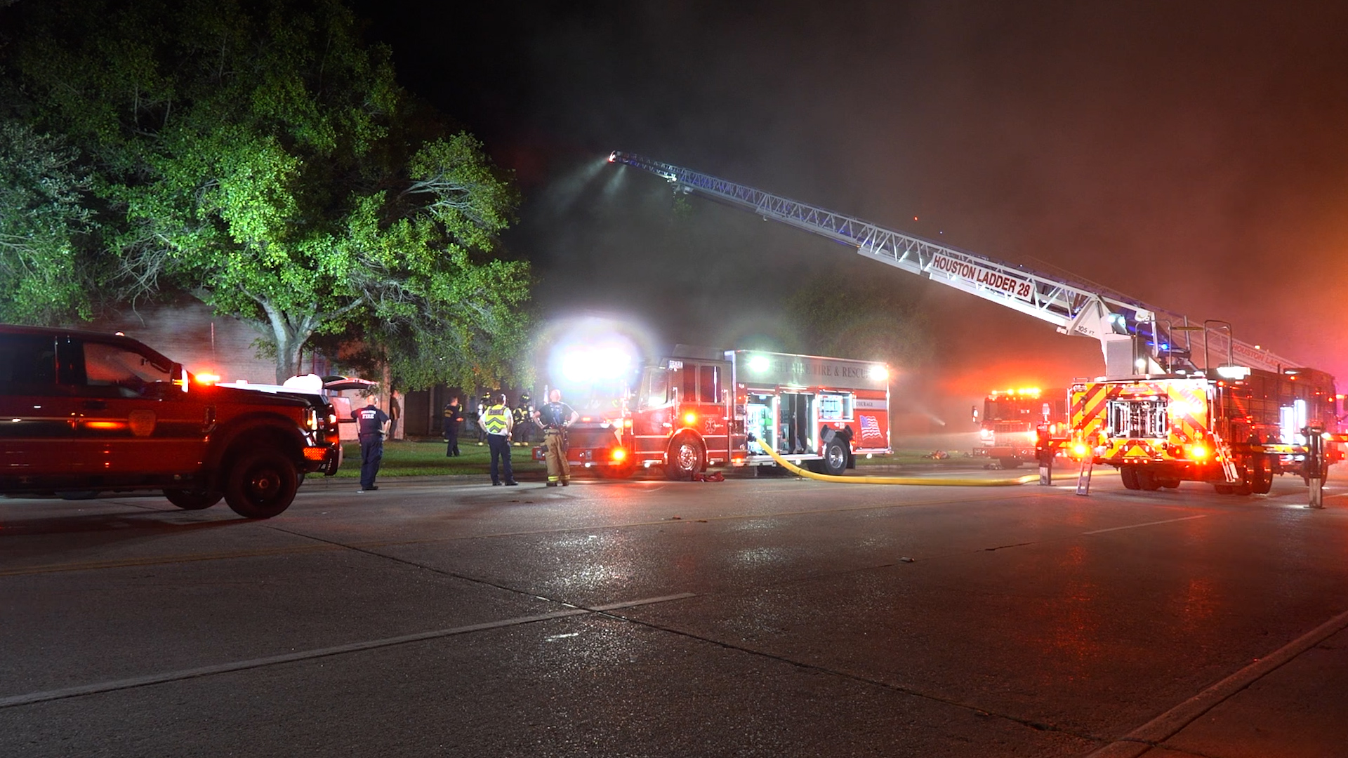 Firefighters and multiple fire trucks operate at night as a long ladder truck extends its aerial ladder toward smoke-filled area with red and blue emergency lights visible.