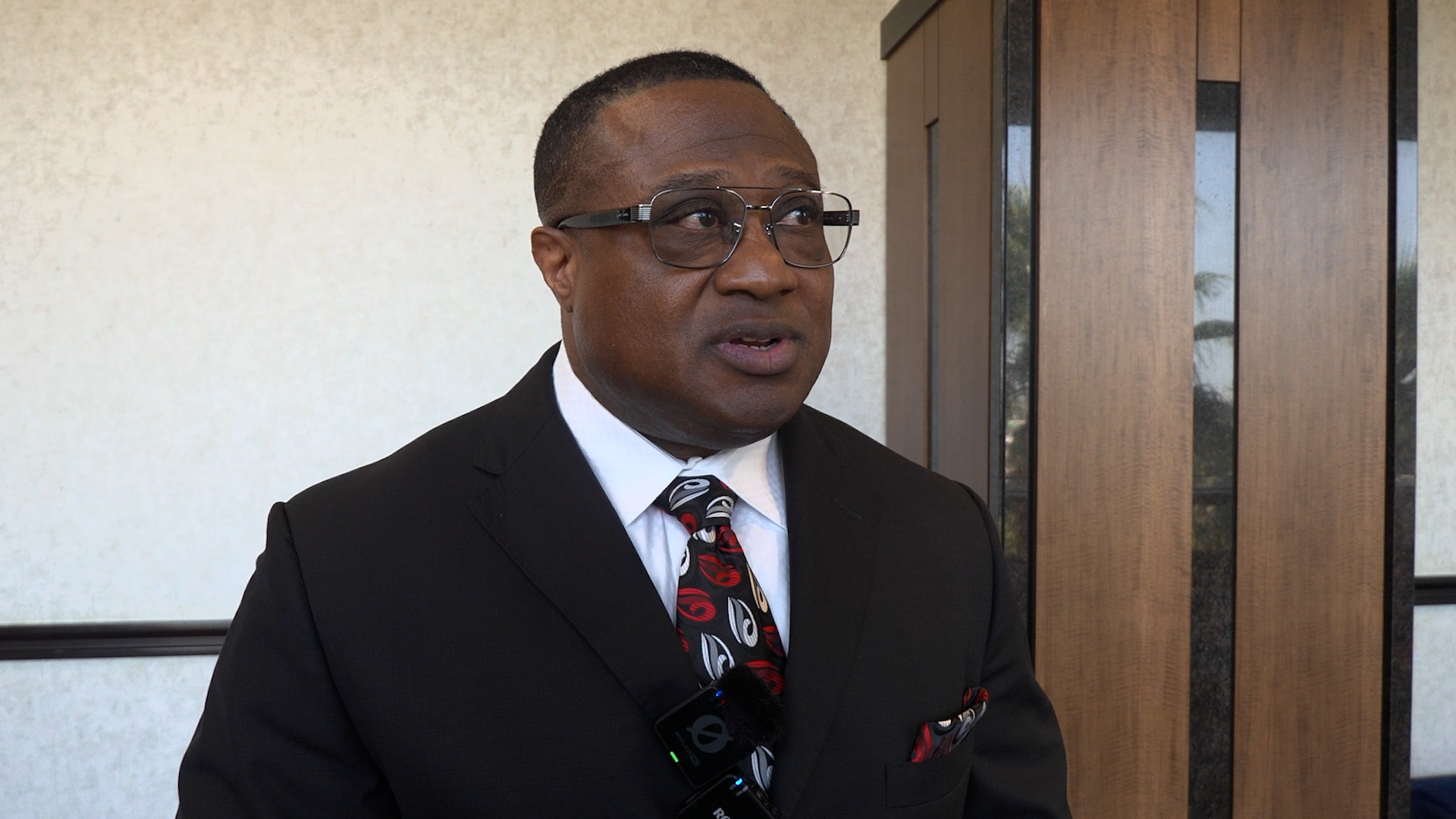 Man in a dark suit and patterned tie being interviewed outdoors, wearing glasses and a lapel microphone clipped to his jacket.