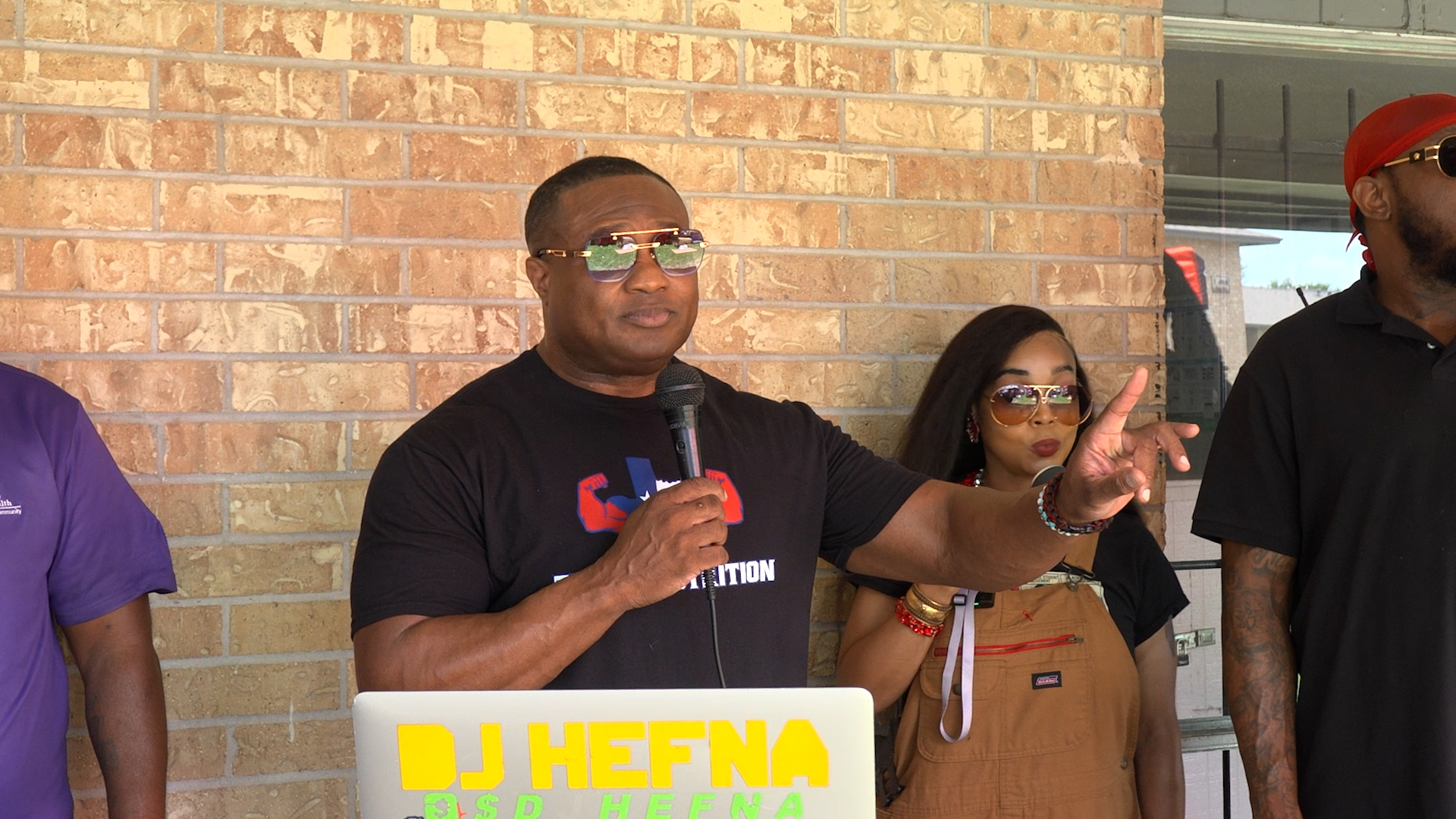 A man in a black shirt speaks into a handheld microphone in front of a brick wall, with a woman and another man visible beside him at a community event.
